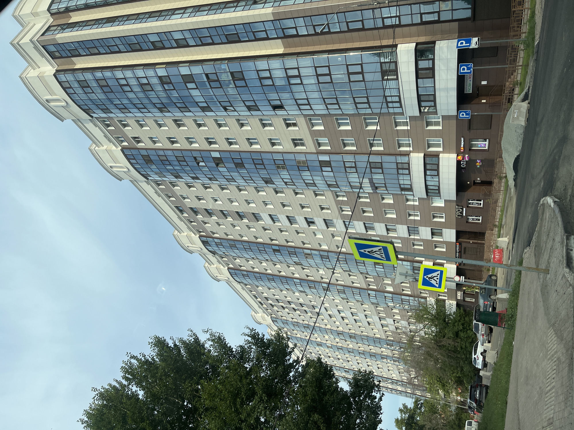The image shows a modern, multi-story apartment building with a facade featuring a mix of brown and light-colored panels, accented by a large number of reflective blue glass windows. The building is curved, extending along the visible street. To the left of the building, a large, leafy green tree is prominent. In the foreground, a paved area with a distinct striped texture leads to a sidewalk. A street with parked cars is visible. Two yellow pedestrian crossing signs with blue pedestrian icons are mounted on poles. There are also several blue parking signs with white 'P' symbols. In front of the building's ground floor, which appears to house commercial establishments, there is a pile of gravel and some construction barriers. The sky above is a pale blue with some faint cloud streaks. The scene is set in Novokuybyshevsk, Russia.