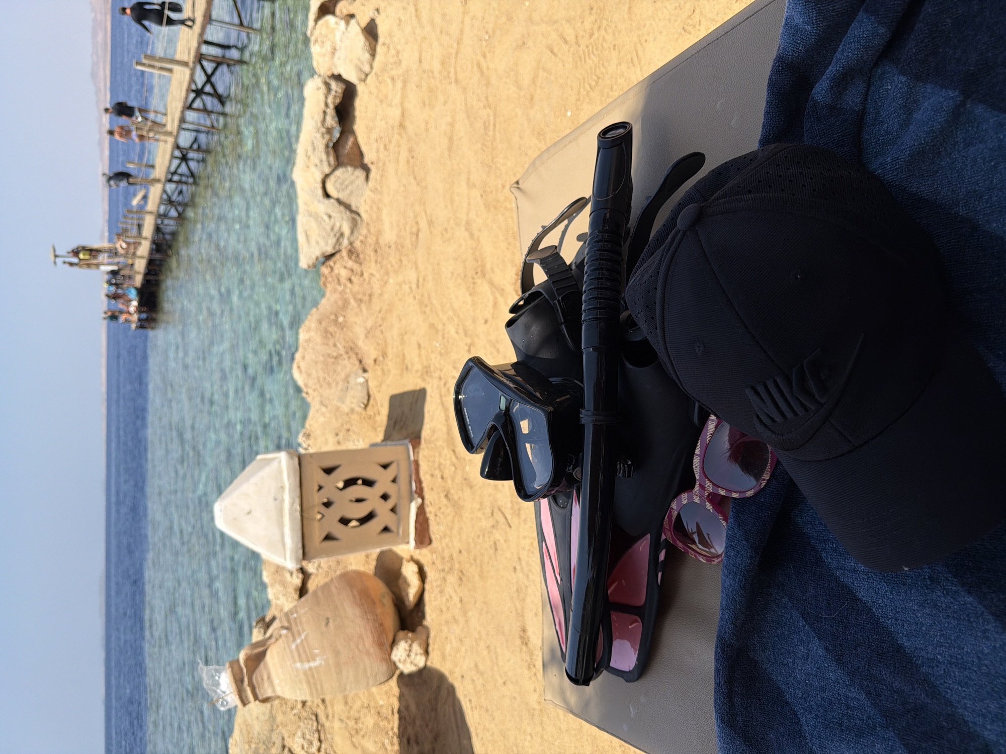 In Sharm El Sheikh, Egypt, a sun-drenched beach scene unfolds with a focus on snorkeling gear resting on a lounge chair. The foreground features a black diving mask and snorkel, paired with pink-accented fins. Adjacent to these are a pair of pink sunglasses with a repeating logo pattern and a black baseball cap. A dark blue towel is draped over the lounge chair, partially obscuring a black cap. In the background, the crystal-clear turquoise waters of the Red Sea extend towards a wooden pier where several individuals are visible, some appearing to be preparing for or returning from diving. The beach itself is sandy, with rocky outcrops and a large terracotta amphora and a decorative lantern placed on the sand. The sky is a clear, bright blue, suggesting a warm and sunny day.