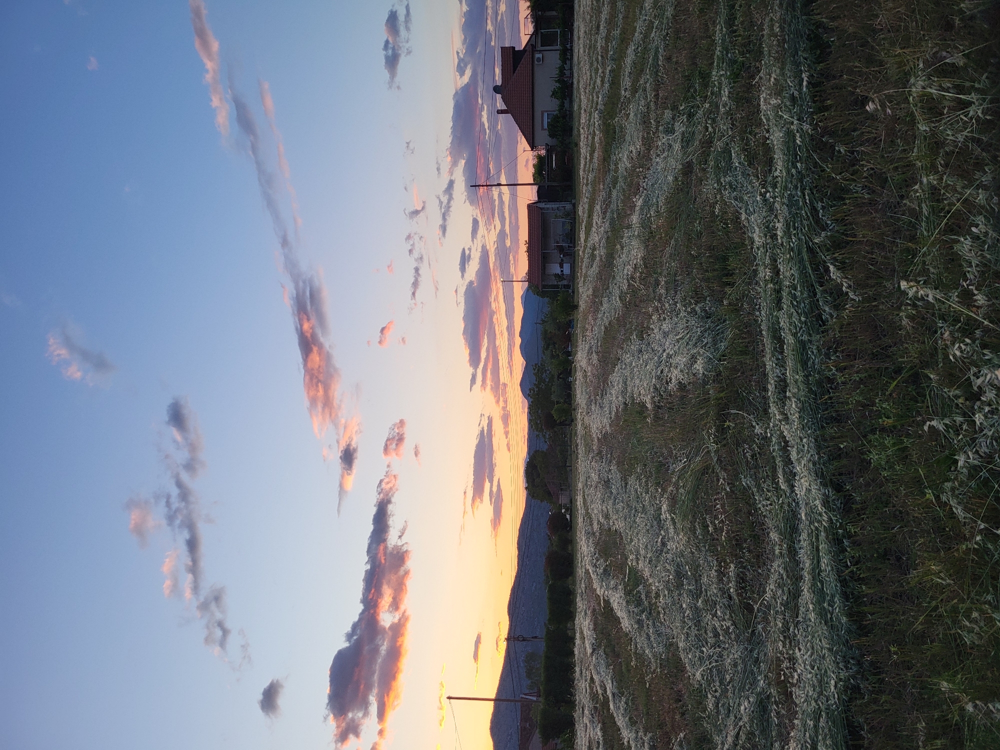 The image depicts a landscape scene near Mahala, Montenegro at sunset. In the foreground, there's a field of cut grass, arranged in rows. Moving towards the middle distance, there are houses and utility poles with wires. In the background, there are hills and a sky with orange and pink clouds. The sky transitions from blue in the upper portion to orange in the lower portion.