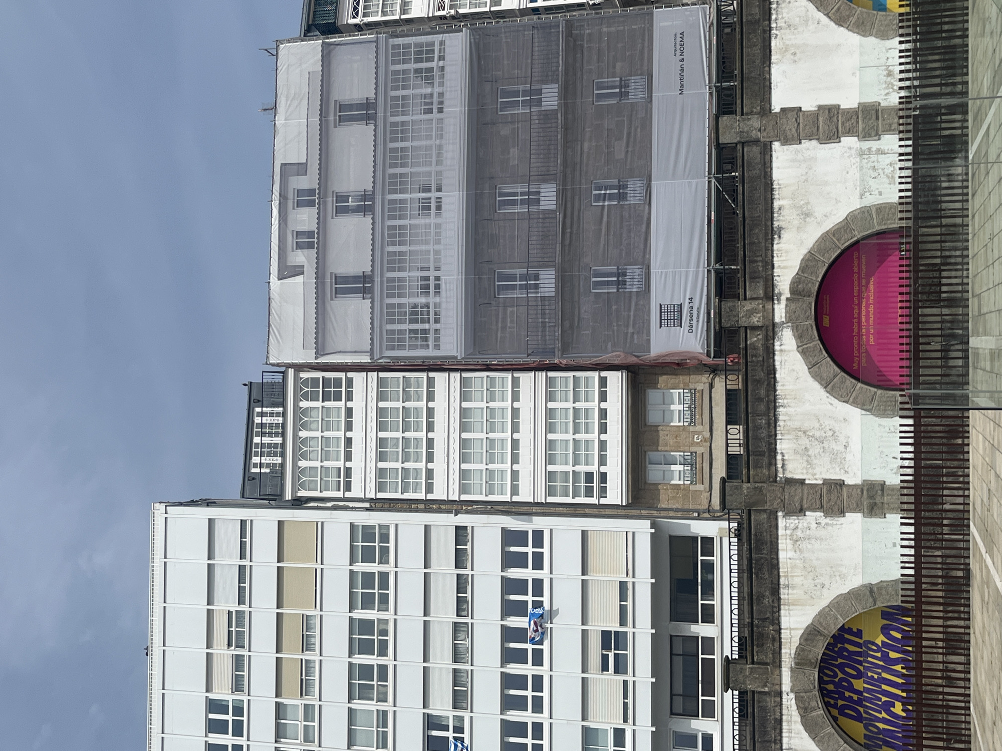 The image shows a street view in A Coruña, Spain, presenting three buildings. The building on the left is modern, tall, and white, with numerous rectangular windows in a grid pattern. There is a blue and white flag visible on one of the windows. The central building is covered in a scrim printed with an image representing its facade, likely for renovation or construction purposes. The building on the right has a stone facade, with two semi-circular alcoves featuring circular advertising posters in a bold color palette of pink and yellow, partially obscured by what appears to be a metal railing or fence. The sky is light blue with some clouds. The OCR in the images describes one of the buildings as the project of the architecture firm Mantinan & Noema, "Darsena 14". Another of the posters refers to sport and movement, promoting an inclusive world.