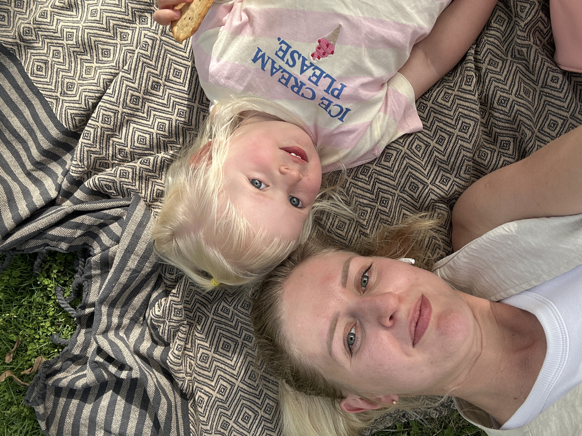 A woman and a young child are lying on a patterned blanket in a grassy area. The child, wearing a striped shirt with "Ice Cream Please" printed on it, holds a cracker. The woman wears a white shirt and has an earbud in her ear. The blanket has a geometric pattern and a striped edge. Some green grass is visible around the edge of the blanket. The photograph appears to be taken outdoors, possibly in a park.