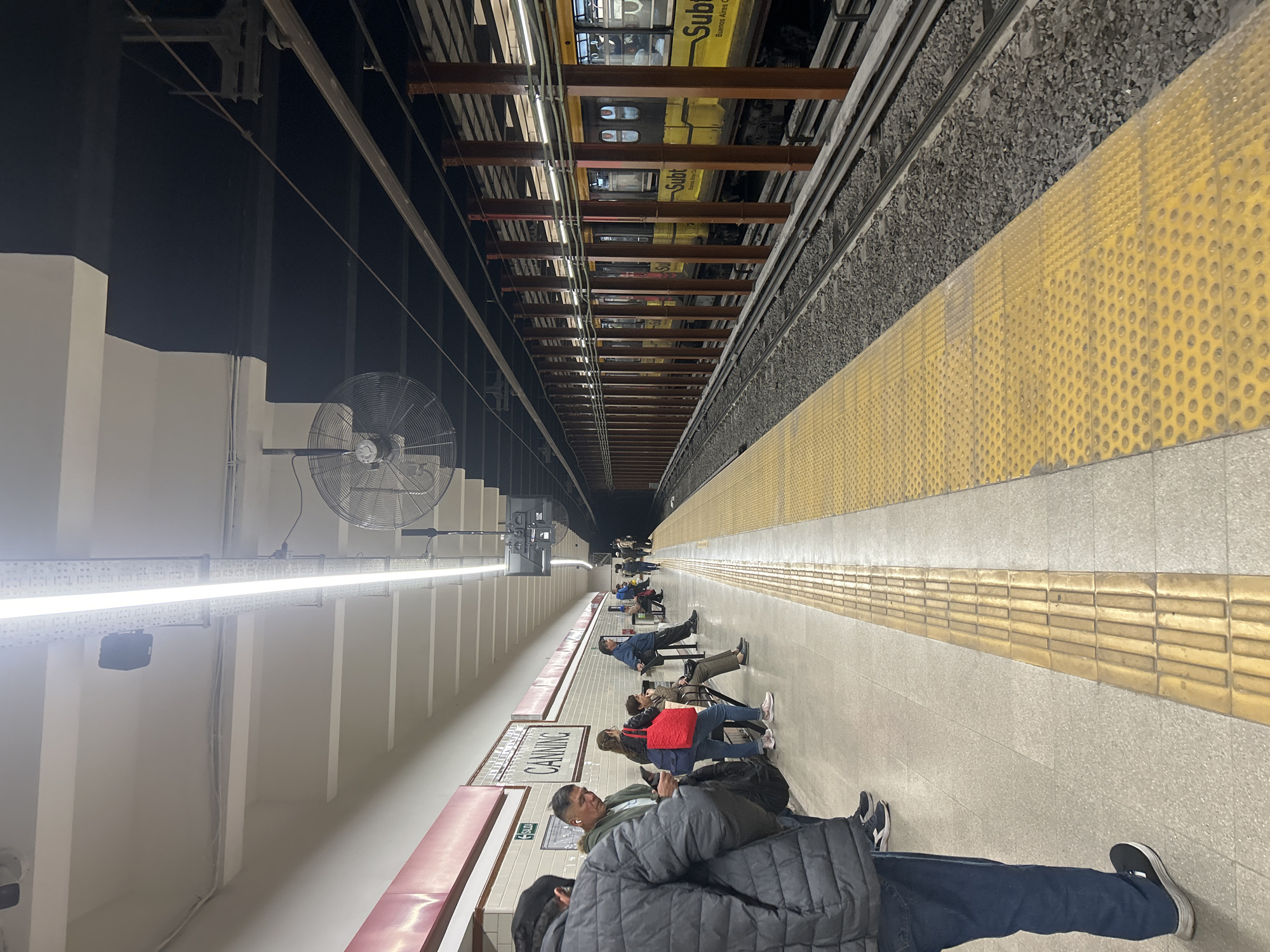This image captures an underground subway station platform, viewed as if correctly oriented despite the original vertical capture. The scene depicts a typical commuter setting in Buenos Aires, Argentina, as evidenced by the "Subte Buenos Aires" text on the train and the station name.
Numerous people are visible along the platform, predominantly seated on red-backed benches, patiently waiting for a train. They appear casually dressed, with some wearing jackets. In the foreground, a person is partially visible, wearing a dark blue puffer jacket and jeans, with wireless earbuds in their ears. Other commuters further down the platform are similarly engaged, some looking at their phones.
The platform itself is made of light-colored concrete, featuring prominent yellow tactile paving in strips and raised dots along the edge, indicating a safety zone. Bright, long linear light fixtures illuminate the platform, casting a stark white light. Large, round industrial fans are mounted on the wall structure, likely for ventilation. Beyond the platform, train tracks laid on grey ballast are visible. A yellow subway train, bearing the inscription "Subte Buenos Aires," is partially seen on the tracks or in an adjacent tunnel. A metallic, latticed overhead structure spans above the tracks. A prominent white sign on the platform wall clearly displays the station name: "CANNING." The overall atmosphere is quiet, characteristic of people waiting during what appears to be either day or night, under artificial lighting.
