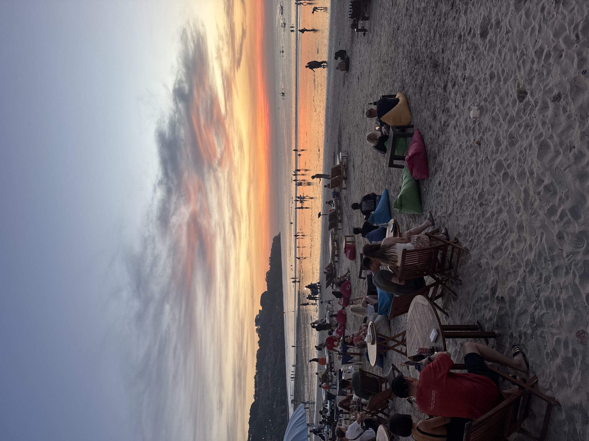 The image depicts a sunset view at a beach in Kuta Selatan, Indonesia. In the foreground, numerous people are sitting on chairs and beanbags at tables on the sand, likely part of a beachside establishment. Many people are facing towards the beach and the sunset. In the middle ground, a wide sandy beach is crowded with silhouetted figures of people walking along the shoreline. In the background is the ocean and the sky, displaying a vivid sunset with a gradient of colors from blue to orange and red, with clouds streaking across the sky.