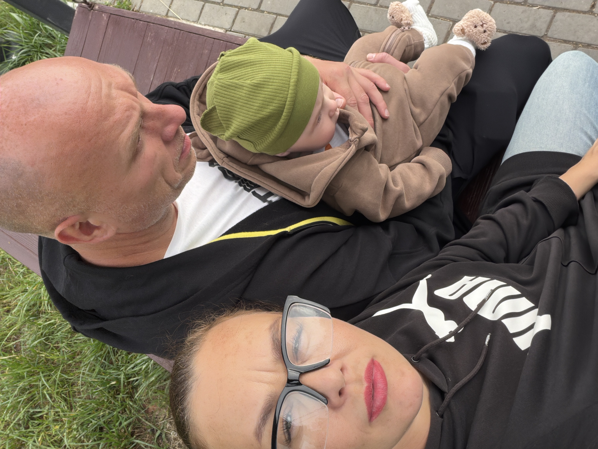 In the city of Chaikovsky, Russia, three individuals are seated on a wooden bench outdoors. On the left, a woman with glasses and red lipstick is looking towards the camera. She is wearing a black Puma hoodie. To her right, a bald man is holding a baby. The baby is wearing a brown onesie and a green hat. The man is wearing a black hoodie over a white t-shirt. The background consists of green grass and a paved walkway. The overall setting suggests a casual outdoor gathering.