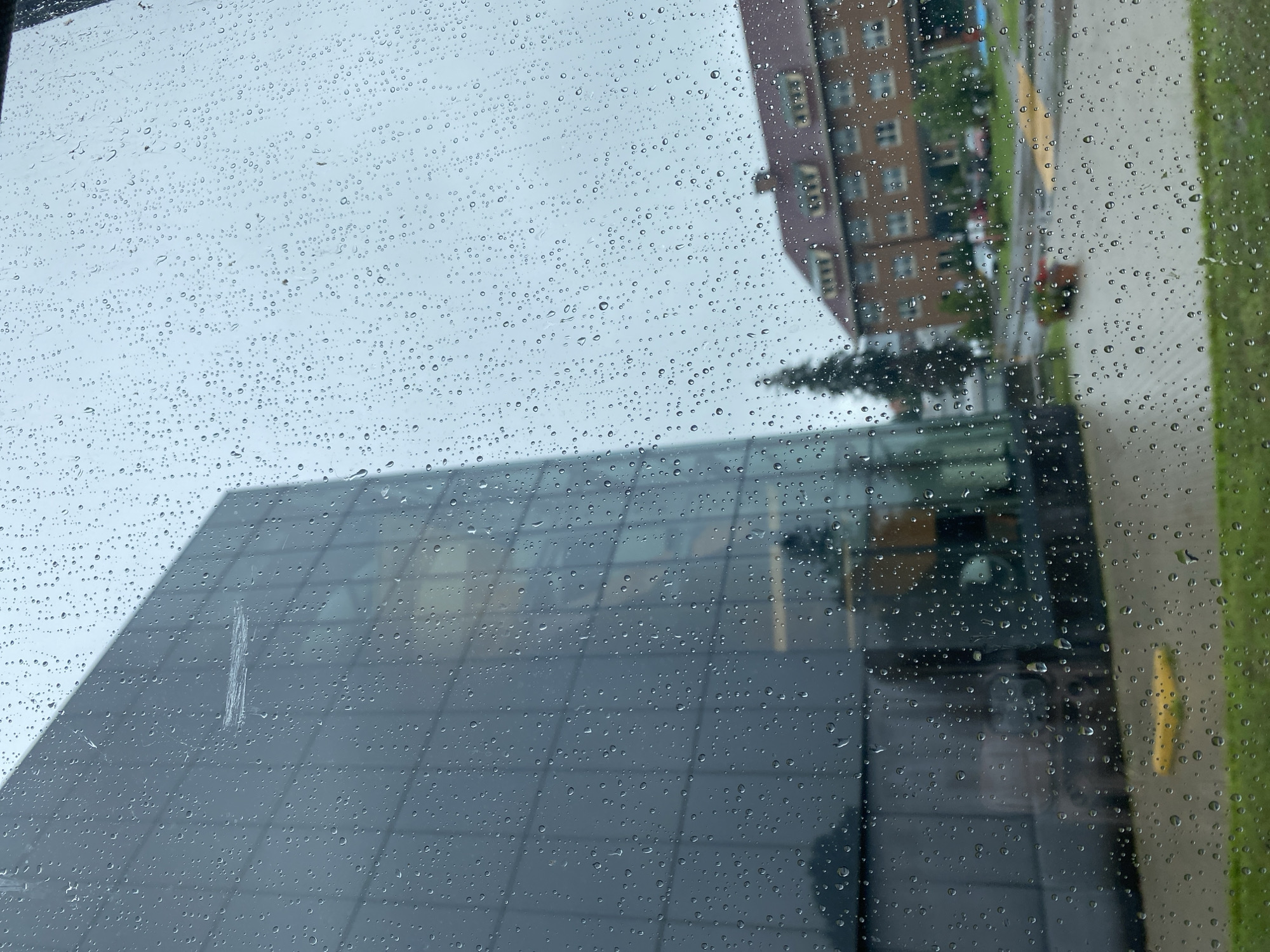 The image depicts a view through a rain-streaked window, likely from inside a building, looking out onto an overcast day in Sainte-Anne-de-Bellevue, Canada. The foreground is dominated by the window's surface covered with numerous water droplets of varying sizes, creating a distorted and textured view of the outside.
On the left, a modern building with a dark, reflective glass facade is prominently visible. Its geometric structure is partially obscured by the rain on the window. A faint scratch or streak is visible on this glass surface, running horizontally. Reflections of interior elements or the sky are subtly visible on the building's glass.
To the right of the modern building, across what appears to be a paved area with some yellow markings and green grass, stands a multi-story brick building with a reddish-brown roof. This building has a more traditional architectural style with visible windows and roof detailing. Between the two buildings, a coniferous tree is discernible. The paved area shows indications of a parking lot or walkway, with a few blurred vehicles and a red planter in the mid-ground. The sky is a uniform gray, consistent with rainy weather. The scene suggests an urban or campus environment.