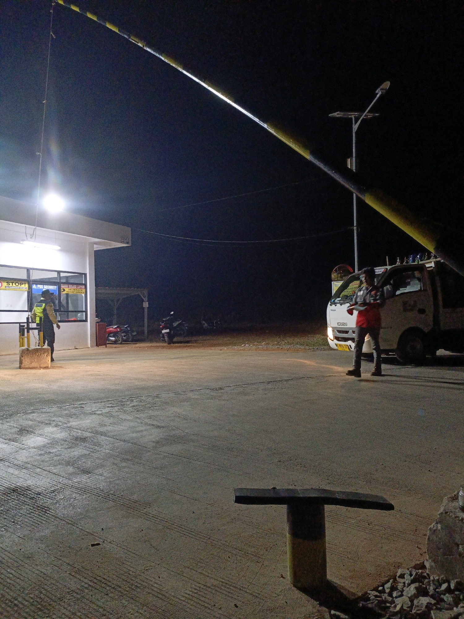 The image depicts a night scene at what appears to be a security post or checkpoint in Jenu, Indonesia. A security guard wearing a yellow vest and dark pants stands near a small building with an illuminated interior. Signs are visible on the building's windows, one of which reads "STOP!". In the background, several motorcycles are parked. A white truck with "ISUZU" visible on the front is also present, and a person in a red vest and dark pants is standing near the truck, holding some papers. A yellow and black striped barrier pole extends across the top of the frame. The ground is paved with concrete, showing tire marks. The overall environment suggests an outdoor location with artificial lighting from the building and a street lamp in the distance.