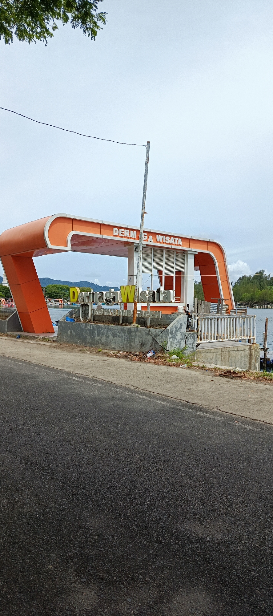 A bright orange, modern archway structure stands beside a waterfront, bearing the Indonesian words "DERMAGA WISATA" in large white letters. Below this, smaller yellow and green letters spell out "DERMAGA WISATA" along with "KOTA BANDA ACEH". A white railing lines the edge of the waterfront. Several boats are visible in the water beyond. The sky is overcast. A dark asphalt road is in the foreground.