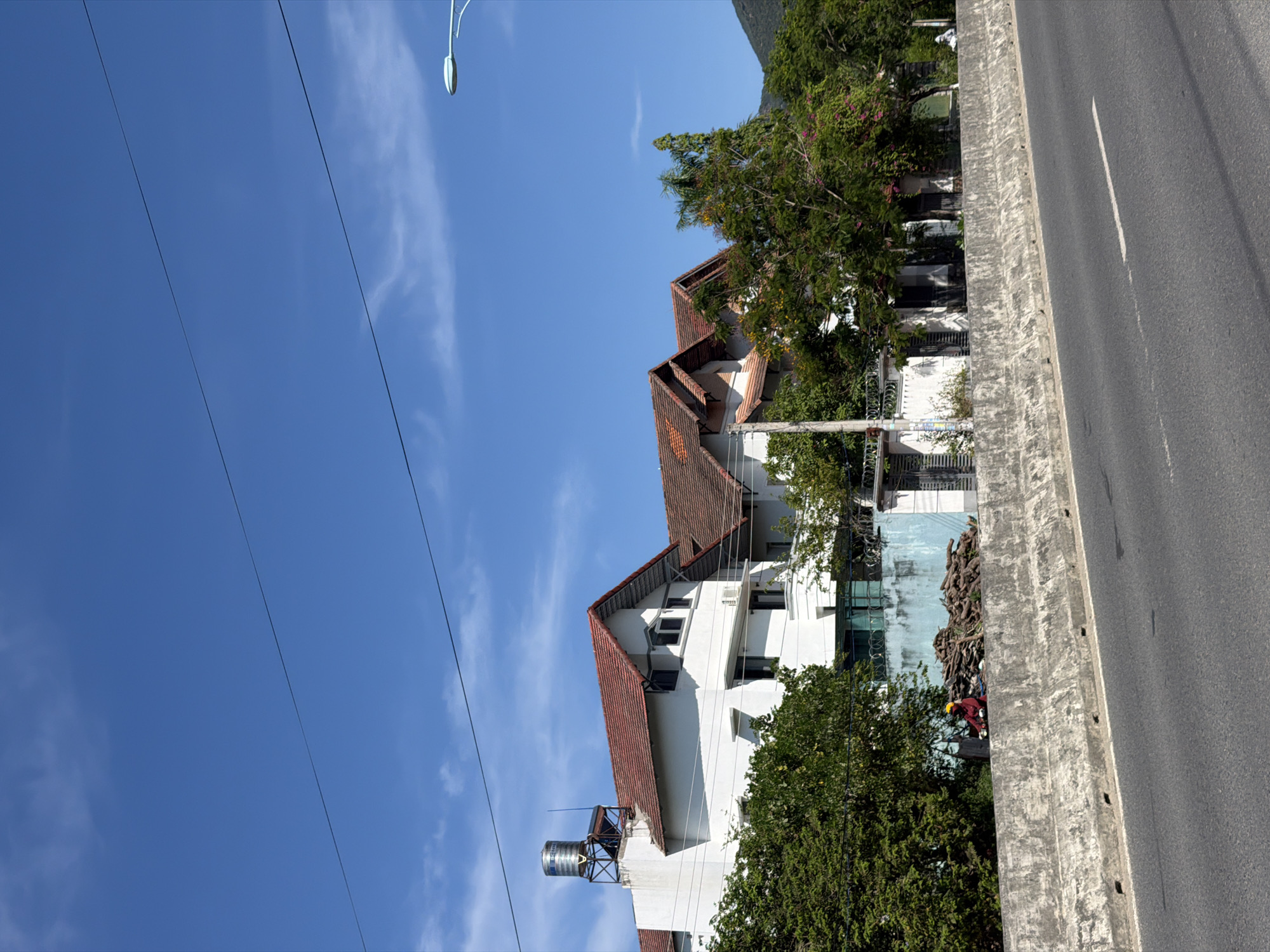 In Nam Nha Trang Ward, Vietnam, a concrete barrier separates a busy road from a line of multi-story white houses with red tiled roofs. The houses are partially obscured by lush green trees. A single person wearing a yellow hard hat and a red shirt sits on a pile of cut logs next to the concrete barrier. The sky above is a clear, bright blue with thin, wispy clouds. A street lamp is visible in the upper right corner of the image.