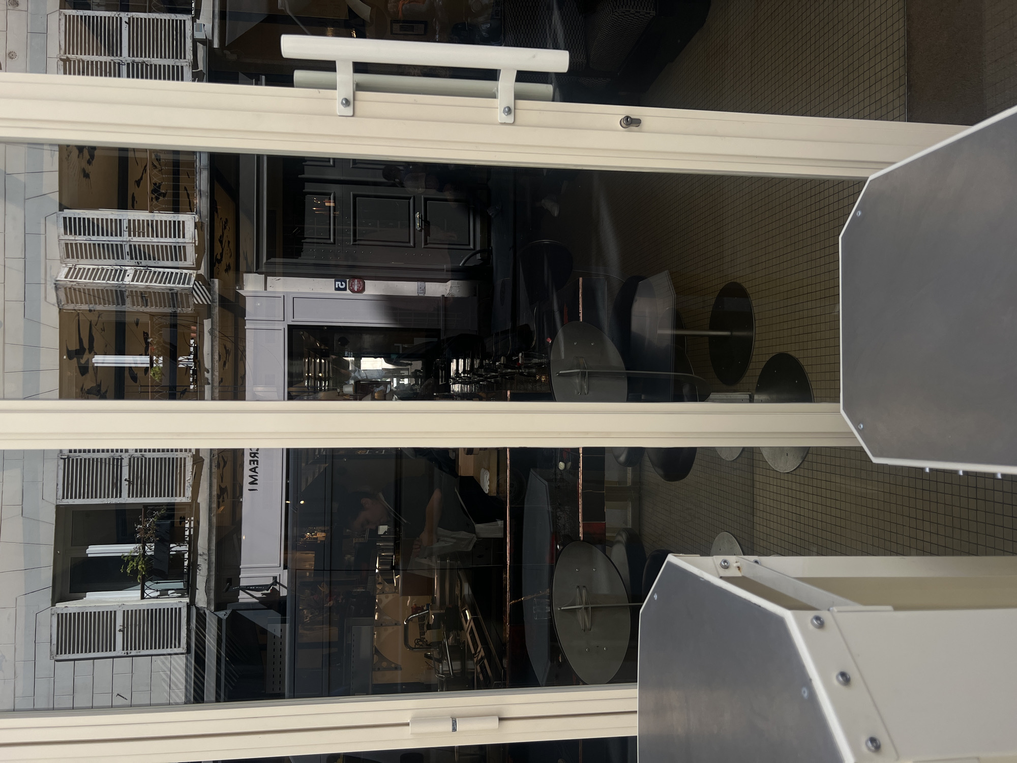 View through a white-framed glass storefront window in Paris, France. Reflected on the exterior is a multi-story building with white shutters. Inside the establishment, an individual wearing a black top appears to be working behind a counter. Additionally, there are black chairs with round bases. The floor is tiled in a grid pattern. A grey service station stands at the foreground.