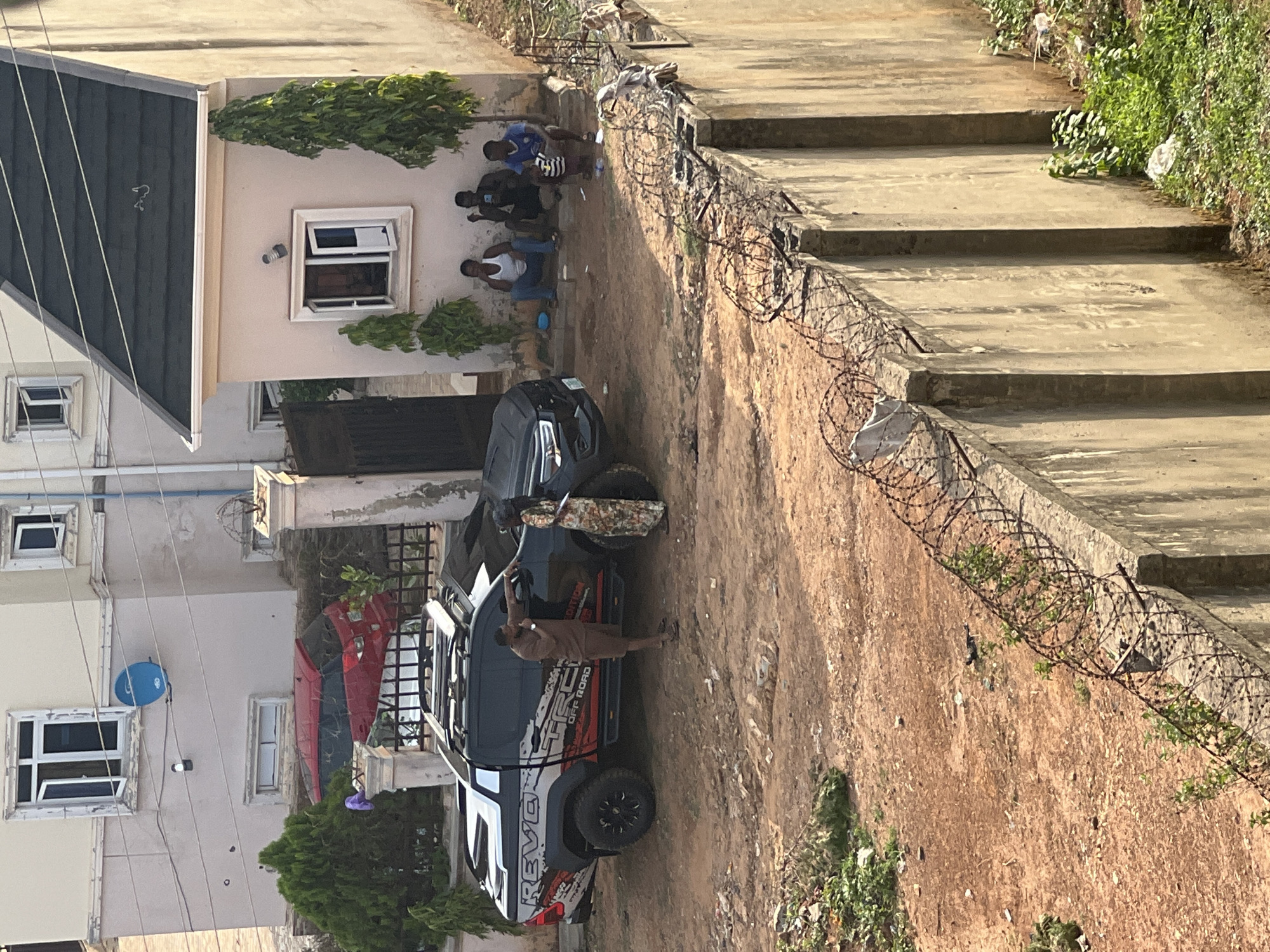 The image shows a scene in Abuja, Nigeria. A white and black pickup truck with the words "REVO OFF ROAD EDITION" on its side is parked on a dirt surface. A man in a brown outfit is standing near the truck's open hood. A red vehicle is partially visible behind the truck. To the left, there is a light-colored building with a blue satellite dish on its side. To the right, a stepped concrete structure is topped with barbed wire. Three people are sitting against the wall of the building, and green plants are visible in the background.