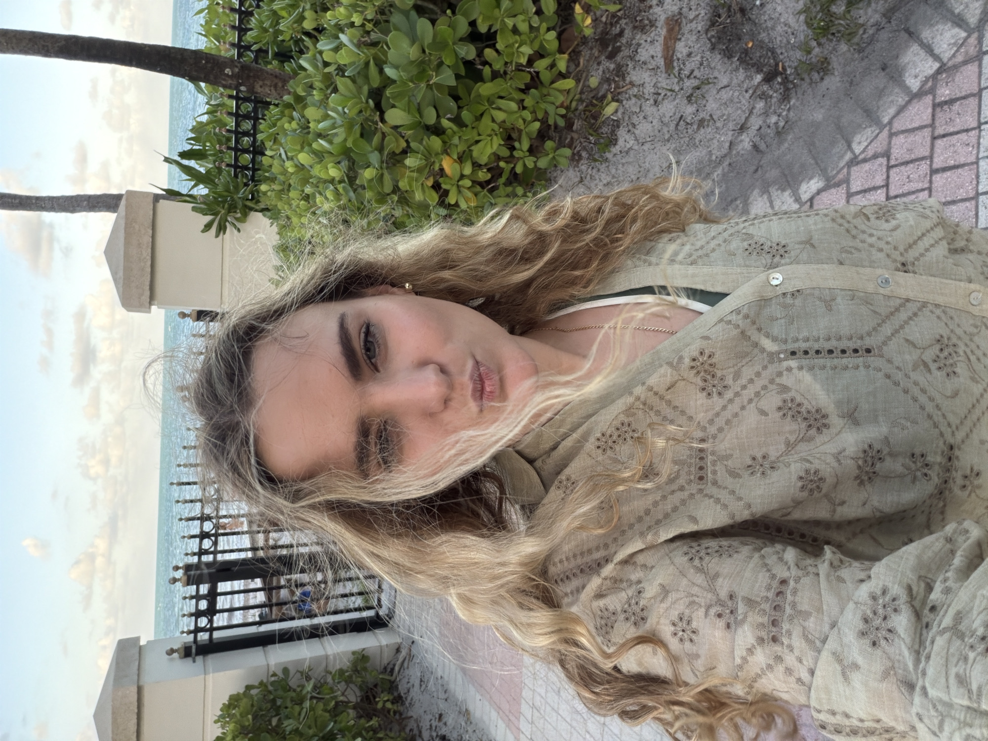 A fair-skinned woman with long blonde curly hair is pictured in a selfie. She is wearing a light tan, embroidered long-sleeved shirt and has a gold chain necklace. The woman has dark eyebrows and is pursing her lips for the photo. The background includes a black wrought iron fence, white concrete pillars, green foliage, a glimpse of the ocean, and a cloudy sky. The location appears to be near a waterfront path in Key Biscayne, United States of America.