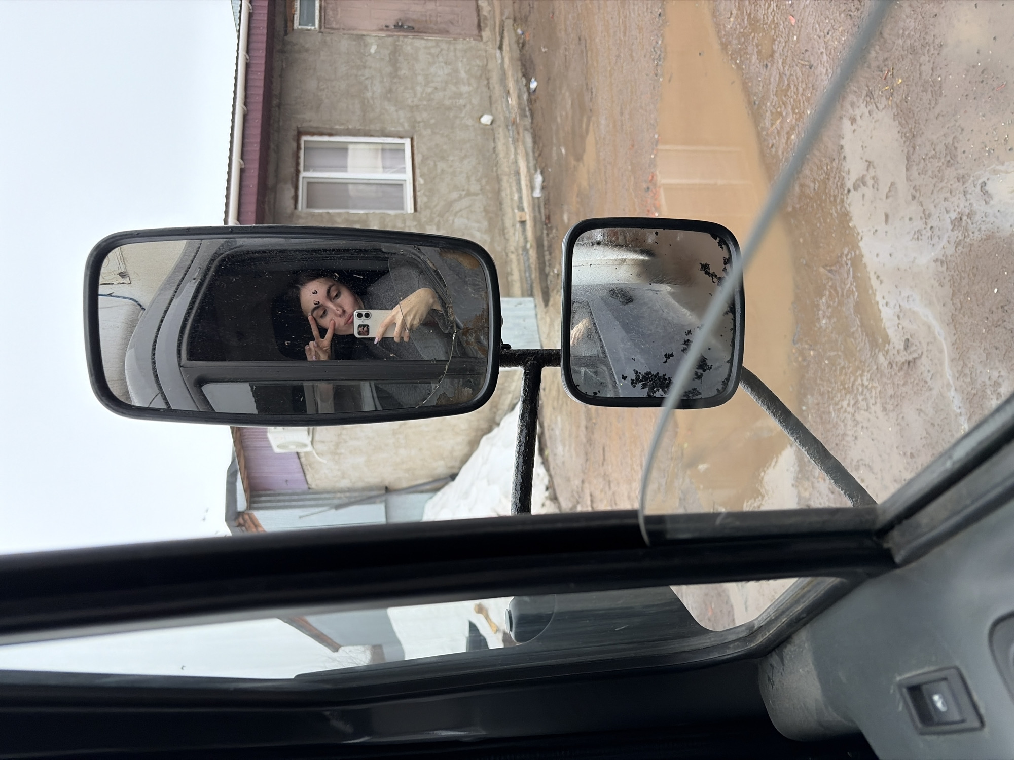 The image depicts an interior view from a vehicle, possibly a truck or a bus, with two side mirrors visible. The left mirror reflects a young woman inside the vehicle, holding a smartphone and posing for a selfie with a peace sign. Her dark hair and a mark on her forehead are visible. The right mirror shows a blurry, rain-splattered street scene. The vehicle appears to be on a muddy road. Outside, a two-story building with a window is visible. Based on visual cues, the location is identified as Izhevsk, Russia. The overall environment suggests a rainy or wet day.