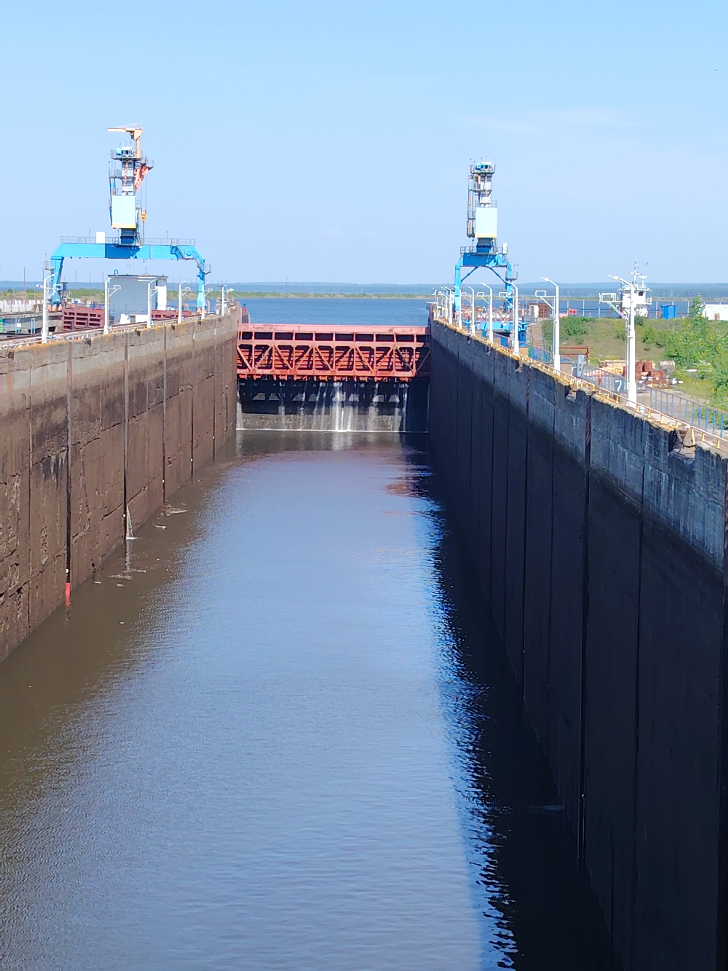 This photograph depicts a large canal lock, likely located in Chaikovsky, Russia, judging by the industrial infrastructure often associated with canal systems in that region. The lock is filled with water, its dark surface reflecting the sky. The massive concrete or steel walls of the lock rise high on either side, showing vertical grooves or seams. A substantial gate, constructed of red metal and featuring a lattice pattern, stands at the far end of the lock. Two towering blue gantry cranes are positioned on either side of the lock, presumably used for maintenance and operation. The sky is clear and blue, suggesting a sunny day. Some vegetation and industrial buildings are visible in the background beyond the far end of the lock. No people are visible in the image.