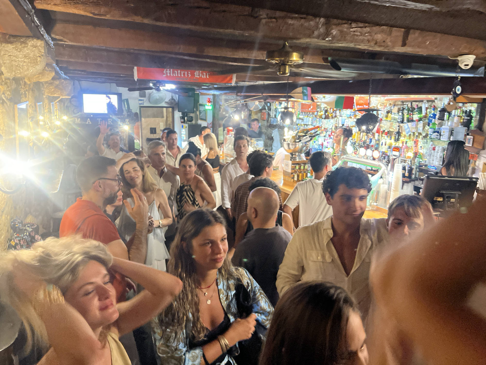 The image captures a crowded interior scene, likely a bar or restaurant, with a lively atmosphere. Several people are visible, some interacting with each other, while others are looking towards the camera or engaged in their own activities. The background features a bar area stocked with bottles, suggesting a place serving drinks. A banner with the text "Matriz Bar" is prominently displayed, identifying the establishment. The overall lighting is dim, with some areas illuminated by artificial lights, creating a warm and inviting ambiance. The presence of a TV screen showing an image also adds to the setting. The crowd appears to be a mix of ages, suggesting a popular spot. The architectural elements, such as the wooden beams and stone walls, contribute to the rustic charm of the location. The city of Caminha, Portugal is known for its historic charm and coastal beauty, and this image seems to reflect a vibrant social scene within the city.