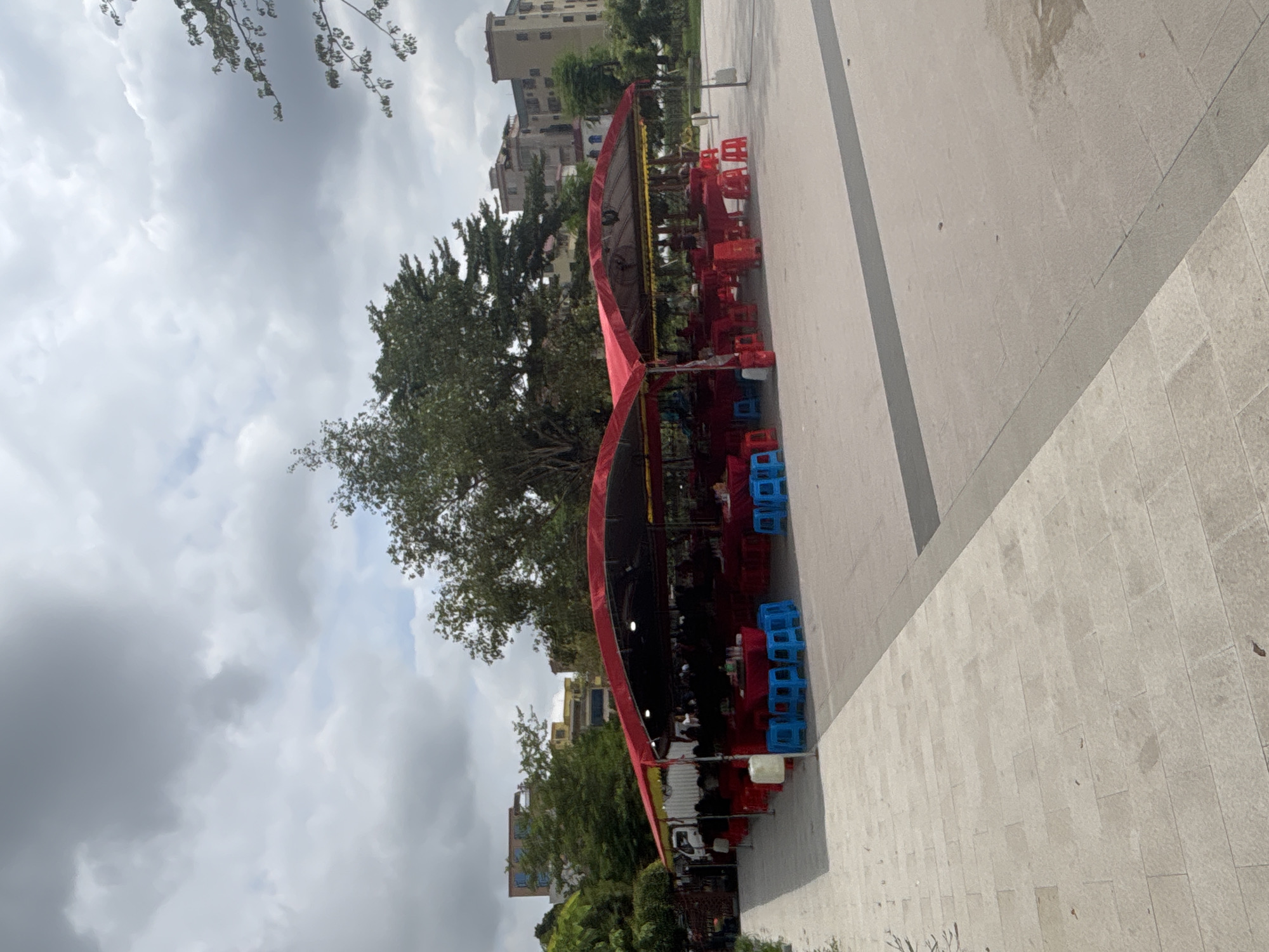 The image shows an outdoor seating area with red tents covering multiple tables and chairs. The area appears to be a food establishment. Several blue plastic stools and red plastic stools are arranged around the tables. In the background, there are trees and buildings, indicating an urban setting. The ground is paved with stone tiles. The sky is cloudy. The scene is located in Nanhai District, China.