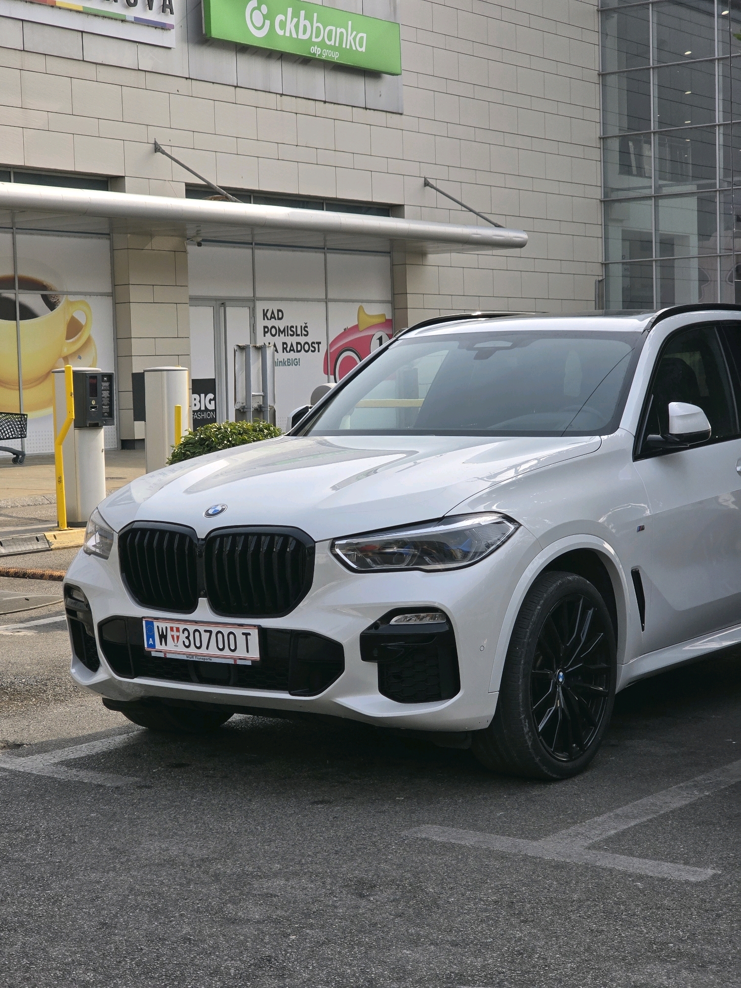 A white BMW X5 is parked in an outdoor parking lot in front of a modern building. The building has a light-colored stone facade and large glass windows. Above the entrance, there are signs with company logos, including "ckbbanka". The BMW has a black grille and black rims. The license plate is visible and reads "W W 30700 T". The parking lot surface is asphalt, and there are white painted parking lines. In the background, there are some green bushes. The weather appears to be overcast. This scene is located at the intersection of Slobodana Jovanovica and Cetinjska Street in Podgorica, Montenegro, in front of the Delta City shopping mall.
