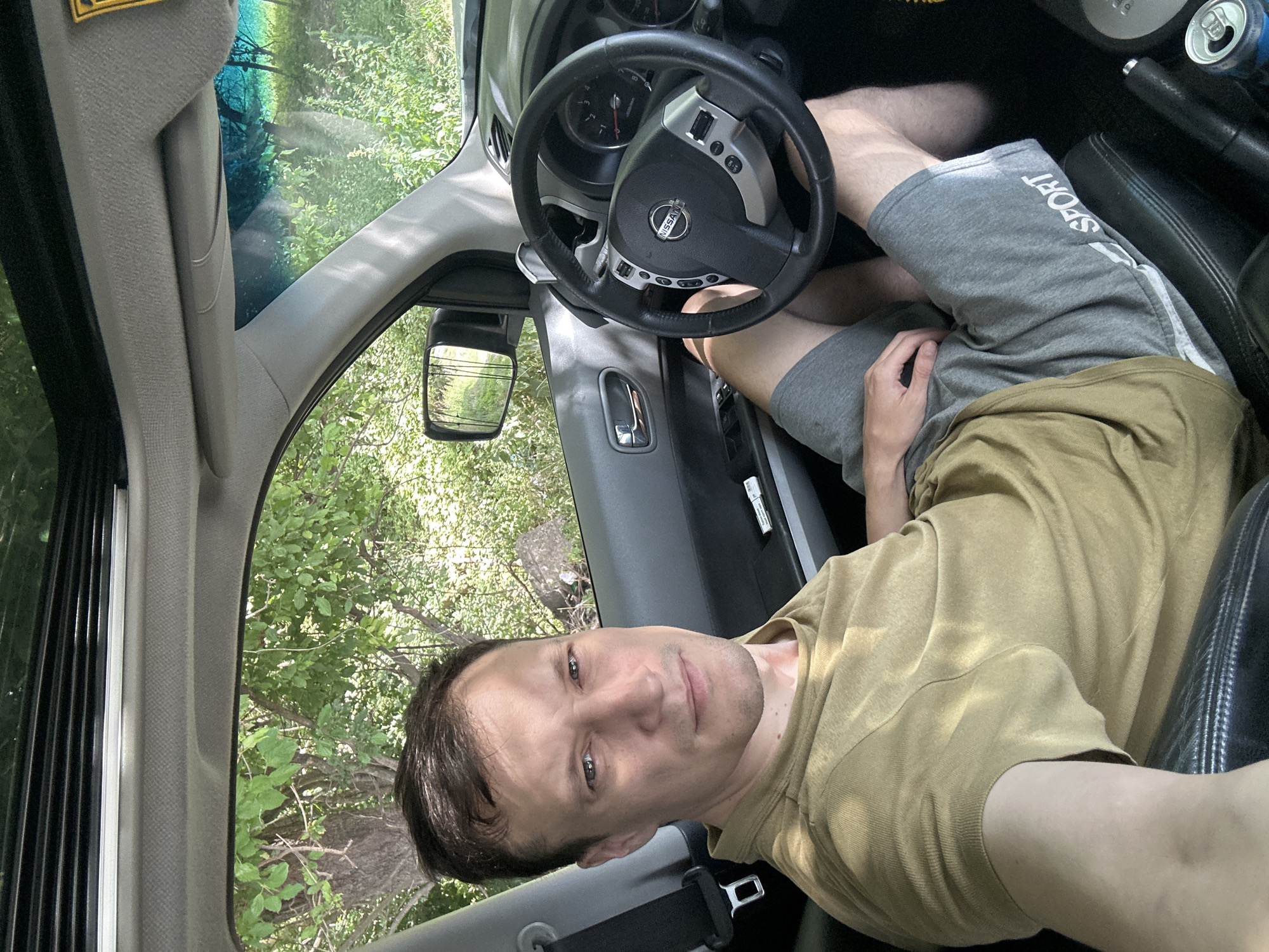 A man is sitting in the driver's seat of a car, looking at the camera. The car is parked outdoors, with trees and foliage visible through the windows and sunroof. The man is wearing a tan t-shirt and gray shorts. The steering wheel and dashboard of the car are visible. A can of soda is in the center console. The scene is likely in Makiv, Ukraine, as indicated by the metadata.