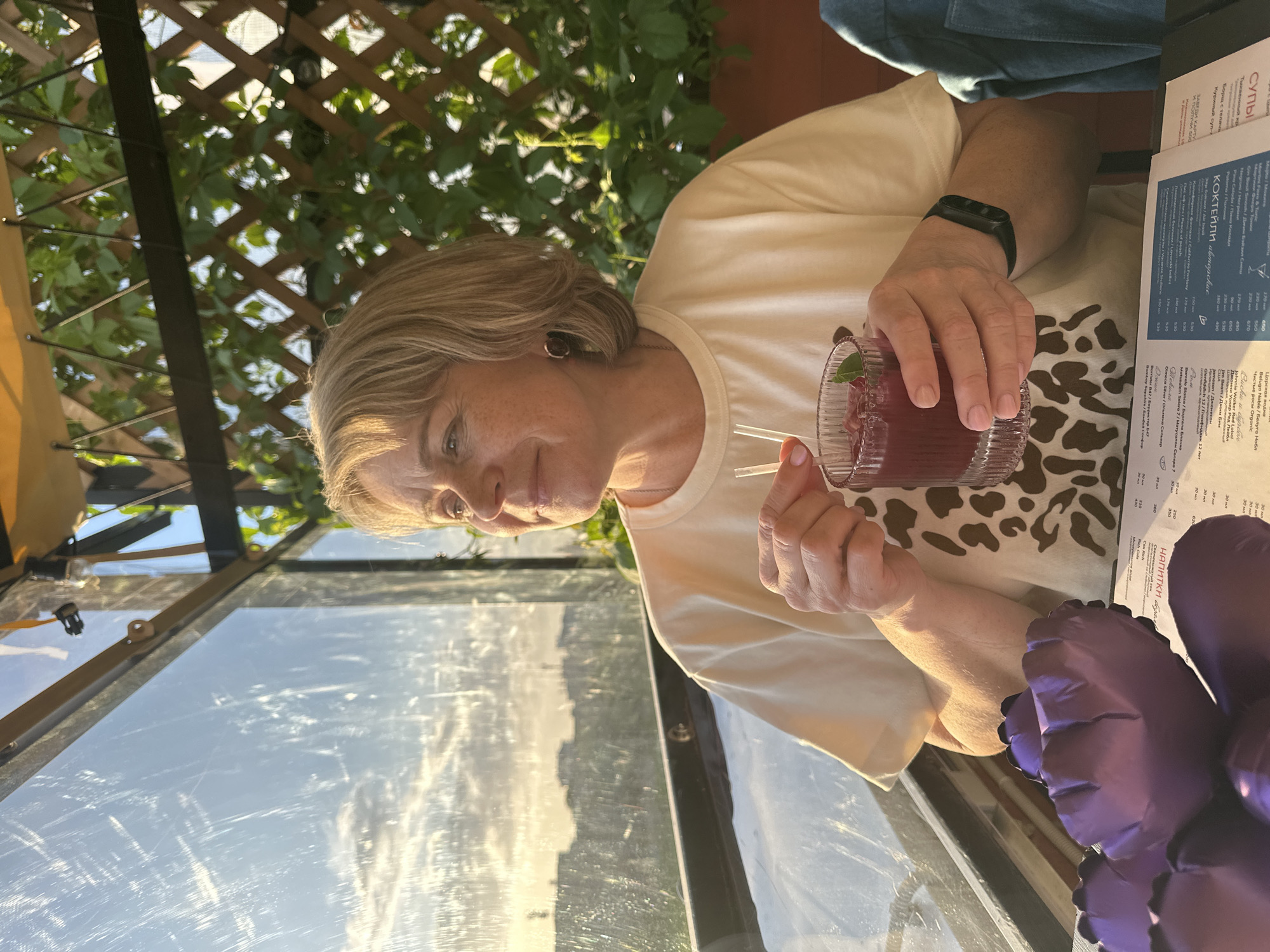 A woman is seated at an outdoor table in what appears to be a restaurant. She is holding a glass of red beverage with two straws. A menu is visible on the table near her right hand. The menu is printed in Russian, and it seems to be a drinks menu with items listed such as “Tsarskaya Vodka," "Martini Fiers & Tonic," and "Pina Colada." The lighting suggests a daytime or early evening setting. The structure surrounding her has a lattice design with green foliage, which may indicate a patio or garden-style area. Outside of the patio, one can see a faint reflection of buildings, suggesting the location is in a city. The OCR from the menu includes "Perm" indicating the city where the photograph was taken.