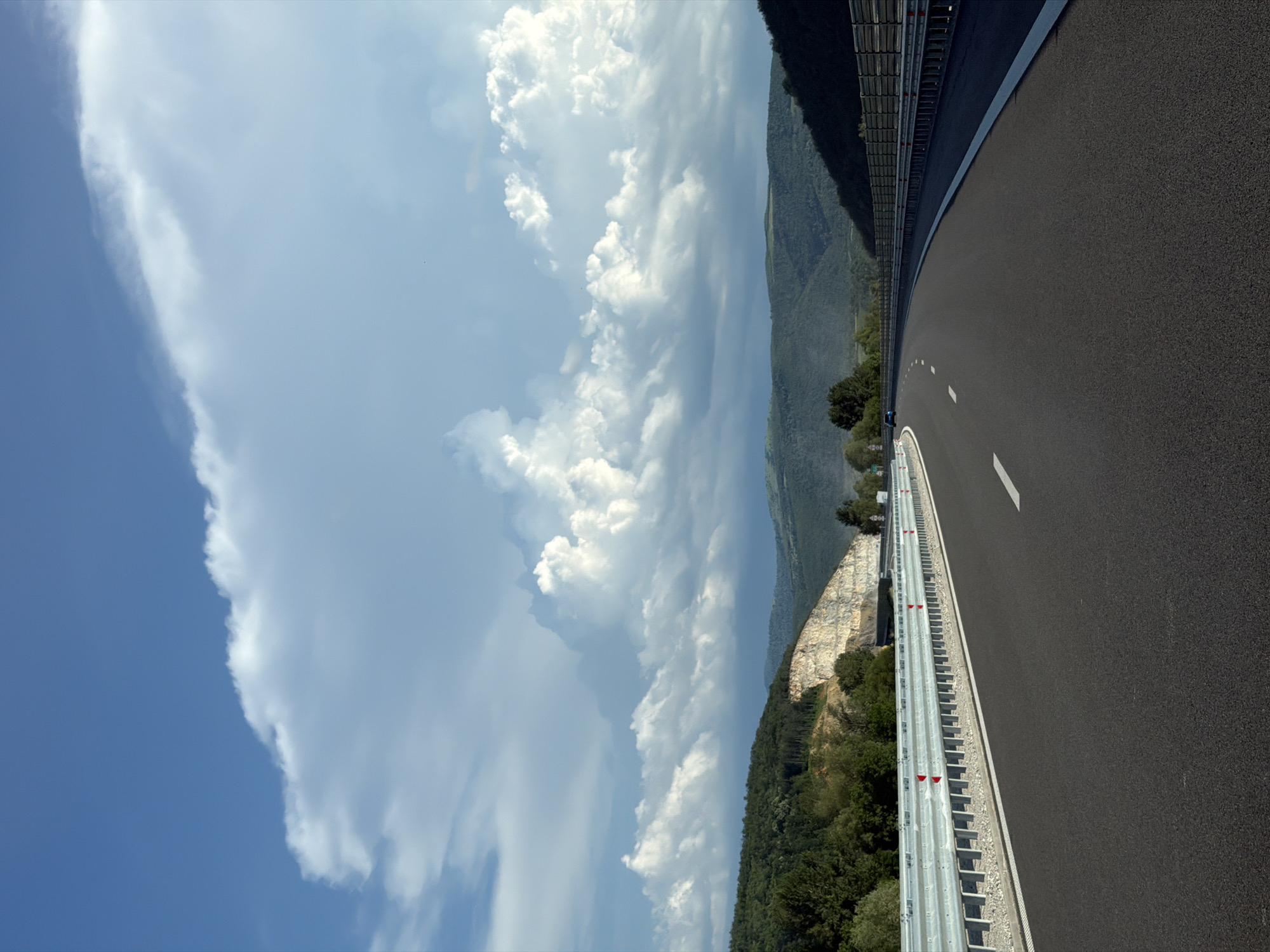 The image depicts a highway scene in Peshterna, Bulgaria, under a dramatic sky filled with large, white, fluffy clouds. The highway itself is paved and dark, with a white dashed line marking the lane. On the left side of the road, a metal guardrail with red reflectors runs along the edge. Beyond the guardrail, a quarry is visible, with layered rock formations and some trees growing on the slopes. The background features rolling hills covered in lush green vegetation, with some darker, more distant mountains. The overall impression is one of a vast, open landscape with a sense of natural beauty and development.