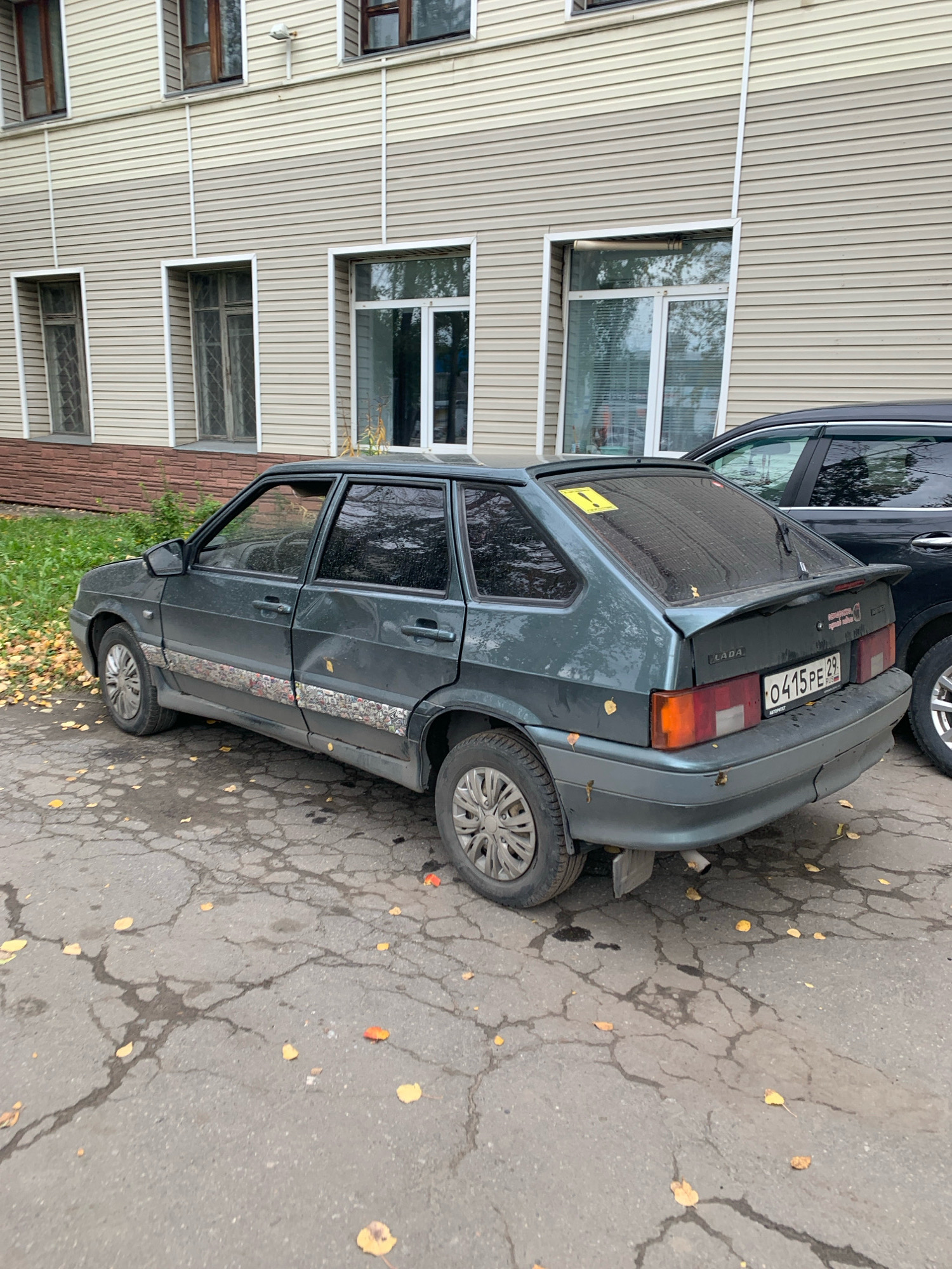 A weathered, dark green Lada hatchback is parked on a cracked asphalt surface covered with fallen yellow and orange leaves. The car has a yellow hazard sticker on its rear window and some visible damage on its side. A dark-colored SUV is parked to the right of the Lada. The background features a building with beige siding and several windows. The scene appears to be an outdoor parking area in Arkhangelsk, Russia.