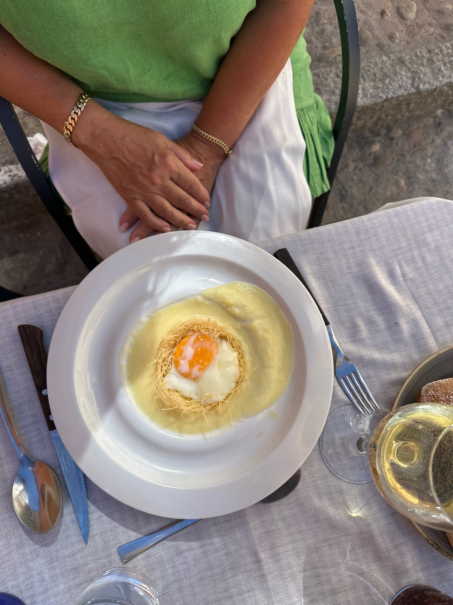 An overhead shot shows a person wearing a green top and white skirt with gold bracelets on each wrist, sitting at a table with a white tablecloth. In front of them is a white plate containing a dish with a fried egg nestled in a nest of shredded dough, surrounded by a creamy sauce. A fork, knife, and spoon are placed on the tablecloth. To the right, a glass of white wine and a plate of bread are visible. The scene appears to be outdoors, possibly at a cafe or restaurant in La Lastrilla, Spain, given the ambient lighting and cobblestone-like background on the right.