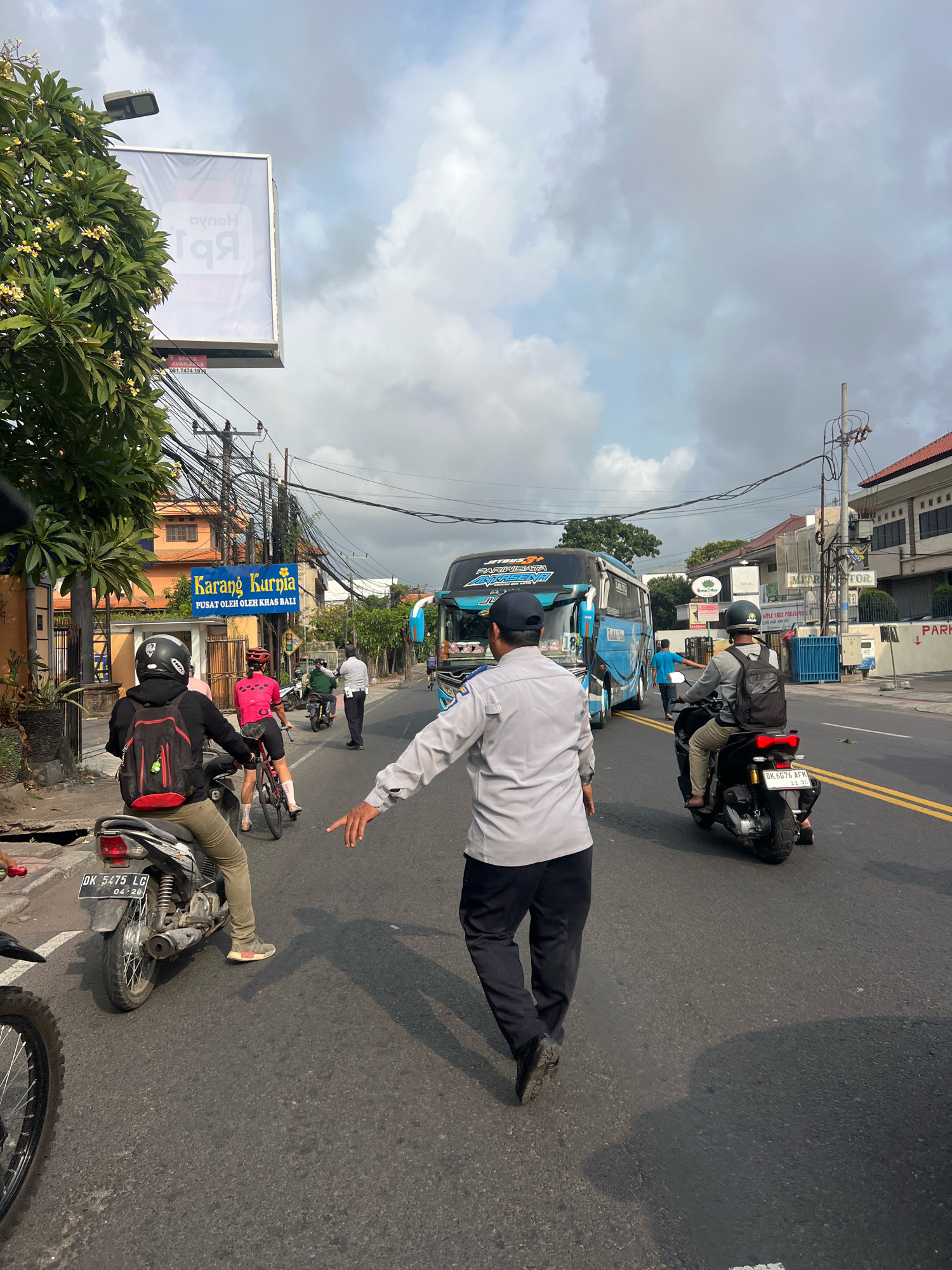 The image shows a street scene in Denpasar, Indonesia. A traffic controller in a light gray uniform and dark pants is gesturing with his right hand towards the right side of the road, indicating traffic direction. He is facing away from the camera and is in the foreground. To his left, a person on a motorbike wearing a black helmet and a black jacket with a red backpack is also facing away from the camera. In the background, a large blue and white bus with "JETBUS3+" branding is visible, traveling on the road. Several other motorbikes and a cyclist in a pink top are also present on the street. The environment is urban, with buildings, power lines, and trees lining the road. A billboard with Indonesian text and a large "Rp" symbol is visible above the street. The sky is partly cloudy, suggesting it could be daytime.