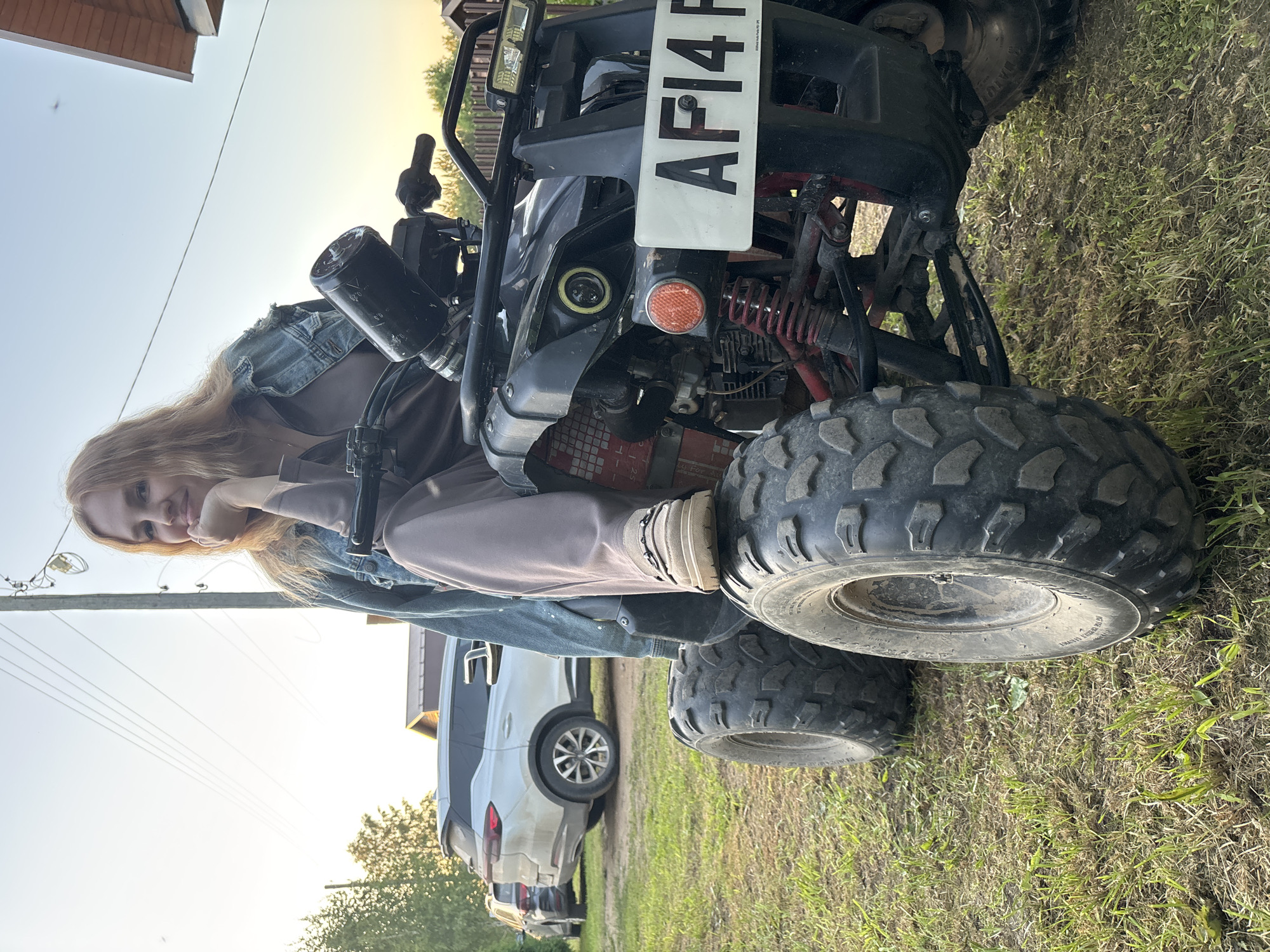 A female subject is seated on a black all-terrain vehicle (ATV) in an outdoor, rural setting in Rozhdestveno, Russia. The subject, an adult female with long, light-colored hair, is wearing a light brown or taupe long-sleeved top and matching pants, with a blue denim jacket draped over her shoulders. Her left hand is positioned under her chin, and she is looking directly at the viewer with a slight smile. Her right leg is bent, with her foot, wearing a light-colored loafer-style shoe with a metallic chain detail, resting on the front left fender of the ATV.
The ATV is equipped with knobby tires, visible red suspension components, and a front-mounted license plate partially displaying "AF14 R". The front of the ATV shows a circular headlight with a yellow halo, an orange turn signal or reflector, and a rectangular LED light bar mounted above. Handlebars and a black cylindrical object, possibly a fluid reservoir, are visible.
In the background, a white SUV is parked facing away from the camera, with another darker vehicle partially visible behind it. A wooden utility pole with multiple power lines extends across the upper left portion of the frame. A light-colored building with a dark roofline is visible in the upper right. The ground is covered with sparse grass and dirt. The scene is illuminated by natural light from a low sun, indicating late afternoon or early evening.