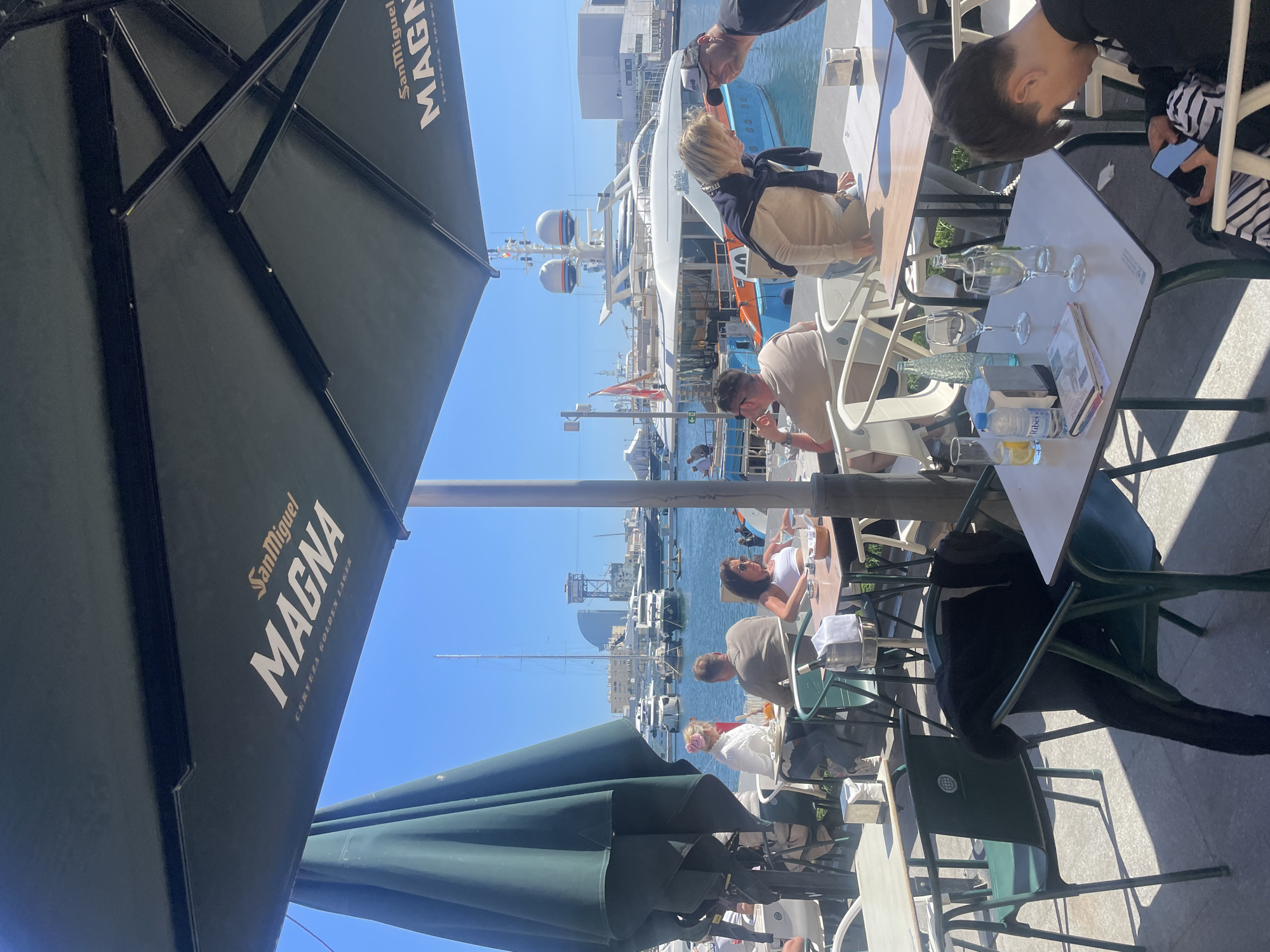 The image shows an outdoor scene with people dining at tables under umbrellas on a sunny day in Barcelona, Spain. The tables are set with glasses, water bottles, and food menus. In the background, there's a view of a marina with boats and buildings, and the clear blue sky. The umbrella in the foreground has the "San Miguel MAGNA" logo. People are seated at tables, enjoying their meals and the view.