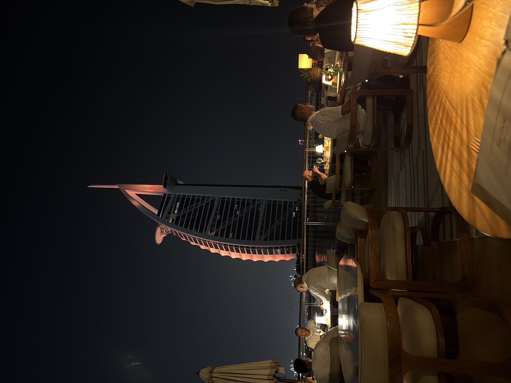 This image was taken at night in Dubai, United Arab Emirates, featuring the Burj Al Arab hotel as seen from an outdoor dining area. The hotel is illuminated with a red glow. The dining area includes wooden tables and chairs, illuminated by lamps. Several people are present, including restaurant staff in white uniforms serving seated patrons.
