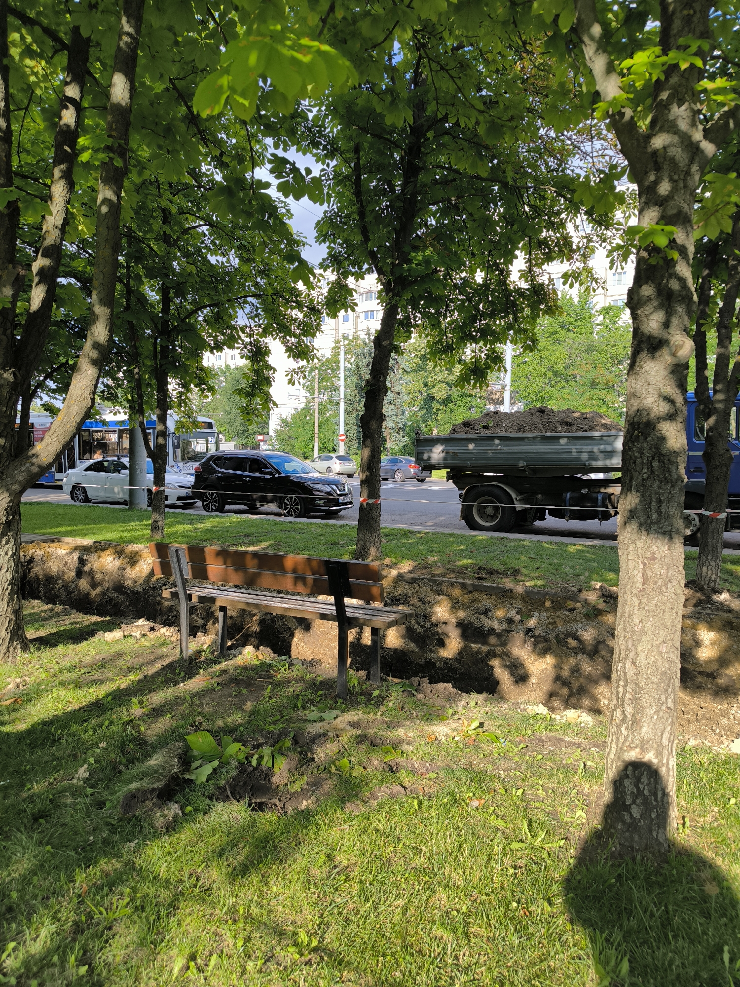The image depicts a sunny day scene in Chișinău, Moldova, likely on a tree-lined street with ongoing construction or landscaping work. In the foreground, a wooden park bench sits on a grassy area. Adjacent to the bench, a trench has been dug in the ground, with piles of dark soil visible along its edges.
Further into the scene, a blue truck with a trailer filled with soil is parked on the road. Several cars, including a black SUV and a white sedan, are also on the road. A public transportation vehicle, possibly a trolleybus, is visible in the background. Trees with lush green leaves are abundant throughout the scene, casting dappled shadows on the ground. The buildings in the background appear to be apartment blocks. Red and white striped caution tape is visible along the edge of the construction area, indicating a safety measure. The overall impression is one of urban activity within a green environment, with elements of both daily life and public works.