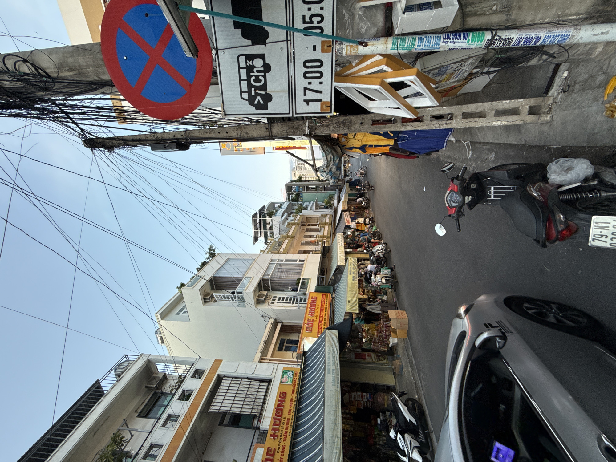 This is a street scene in Nha Trang, Vietnam, likely in the early evening or late afternoon. The sky is clear and a pale blue. The street is lined with multi-story buildings, some appearing to be residential or commercial establishments with awnings and storefronts. There are many overhead electrical wires creating a dense network.
In the foreground, a silver car is partially visible on the left side of the frame. A red and black motorcycle is parked on the right side, with a plastic bag draped over its rear seat. Further down the street, several other motorcycles are parked and some people are walking or standing near the storefronts.
A prominent traffic sign on a pole on the right displays a red circle with a diagonal blue cross, indicating "no stopping." Below this, another sign shows a silhouette of a bus with ">7 Chỗ" (which translates to ">7 seats" or a bus of more than 7 seats) and a time range of "17:00 - 05:00". This suggests a restriction on larger vehicles during specific hours.
The buildings show a mix of architectural styles, with balconies and windows. Some storefronts display signs in Vietnamese, such as "PHUC HUONG," indicating businesses selling goods. The overall impression is of a busy urban street with typical Vietnamese city elements.