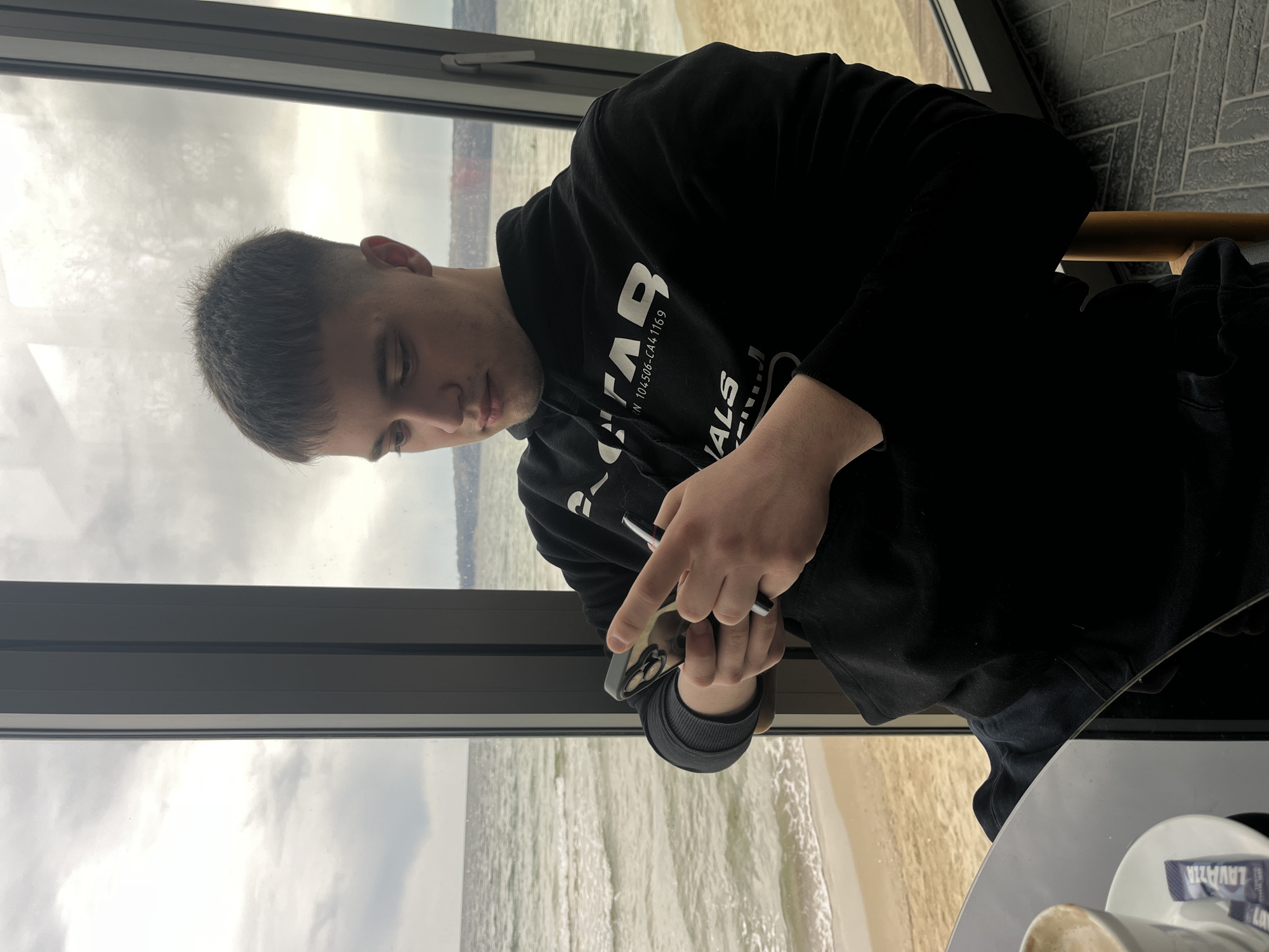 A young adult male is seated inside a building, possibly a cafe or restaurant, looking at a smartphone he is holding in his hands. The man is wearing a black hooded sweatshirt. The background features a large window overlooking the beach and the Black Sea, likely in Varna, Bulgaria, based on the visible coastline and the brand of coffee cup visible on the table. The table is circular and white with a coffee cup on top. The weather outside appears cloudy.