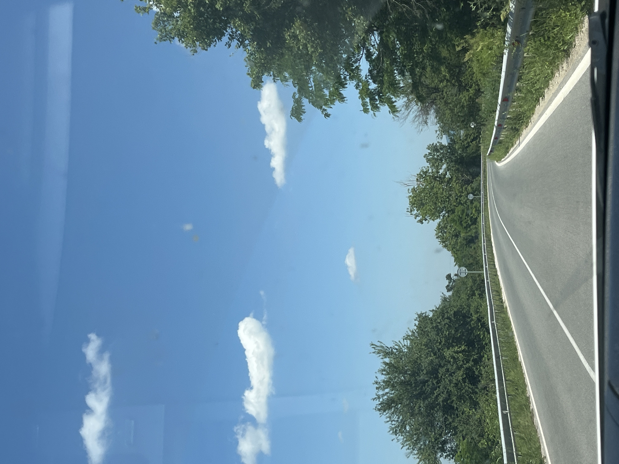 An eye-level, wide shot captures a paved road curving through a rural landscape under a bright blue sky dotted with white clouds. Guardrails line both sides of the road, and dense green foliage and trees flank the roadside, with some trees closer to the viewer on the right and left. The road appears to be in good condition with clear white lane markings. The scene suggests a drive through a natural environment on a sunny day.