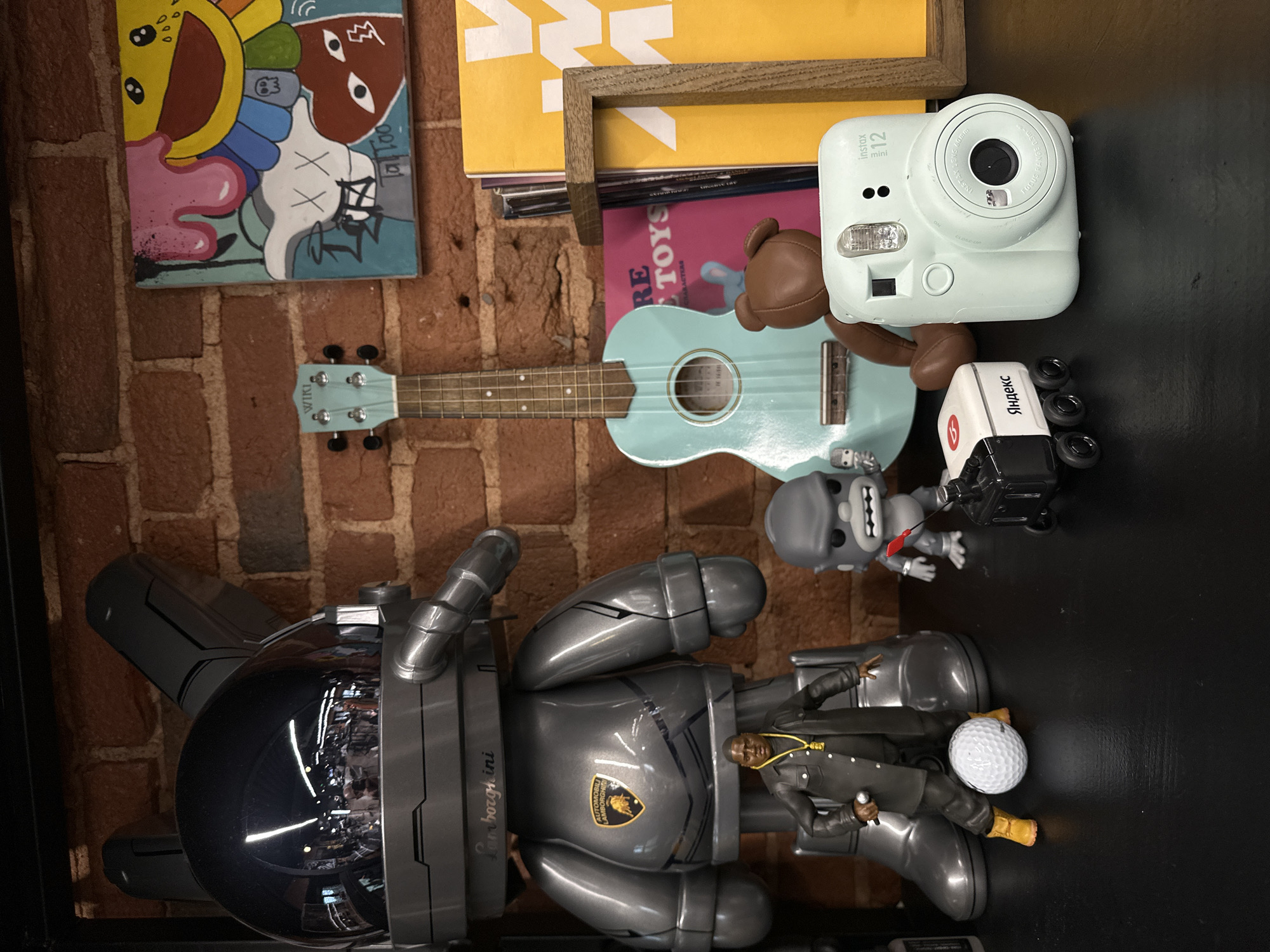 The image displays an eclectic arrangement of collectibles and decorative items, likely positioned on a shelf or tabletop against a warm brick wall backdrop. The collection blends pop culture, branded merchandise, and art.
In the foreground, a large, dark gray robotic-style figure, possibly an art toy like a Bearbrick, is prominent. It features the "Automobili Lamborghini" logo and "Lamborghini" text. At its base, a smaller figurine of a person in a black coat and gold chain, holding a microphone, is placed next to a white golf ball.
To the right, a light blue ukulele with "WIKI" printed on its headstock rests against the brick wall. Above it, a colorful painting or print is visible, featuring cartoonish elements reminiscent of Kaws' "XX" motif and Takashi Murakami's smiling flower. The text "XX" and "Tal Too" are legible on the artwork.
Further right, a light blue Fujifilm Instax Mini 12 instant camera is displayed, bearing "instax mini 12", "INSTAX LENS 60mm", and "FOCUS RANGE 0.3m" on its surface. Beside the camera are a small brown teddy bear figure and a distinctive white robotic toy with "Яндекс" (Yandex) written in Cyrillic, a key indicator for the Moscow, Russia location. Below these, a gray gorilla-like figurine with prominent teeth holds a black accessory.
Stacked books or magazines are visible behind the ukulele, with one clearly labeled "RE TOYS" and "CHARACTERS." The scene presents a curated display of diverse interests, suggesting a personal collection, a retail setting for unique items, or an office space in Moscow.