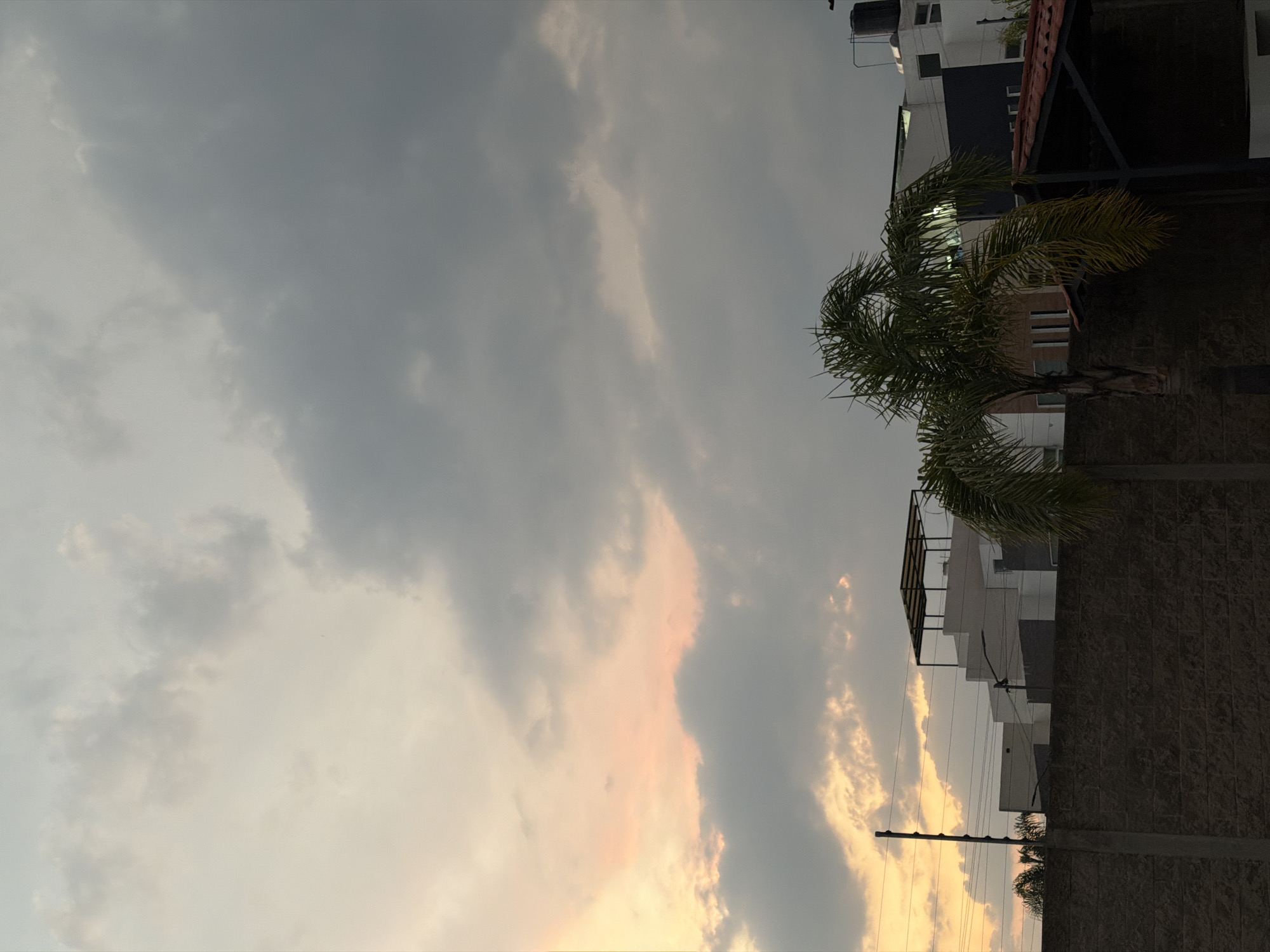 The image, taken in San Juan Cuautlancingo, Mexico, shows a sky filled with grey clouds, tinged with orange from the setting sun. A palm tree is visible against a building with a dark brick or stone wall. The building appears to have multiple levels. Power lines are strung in front of the building.