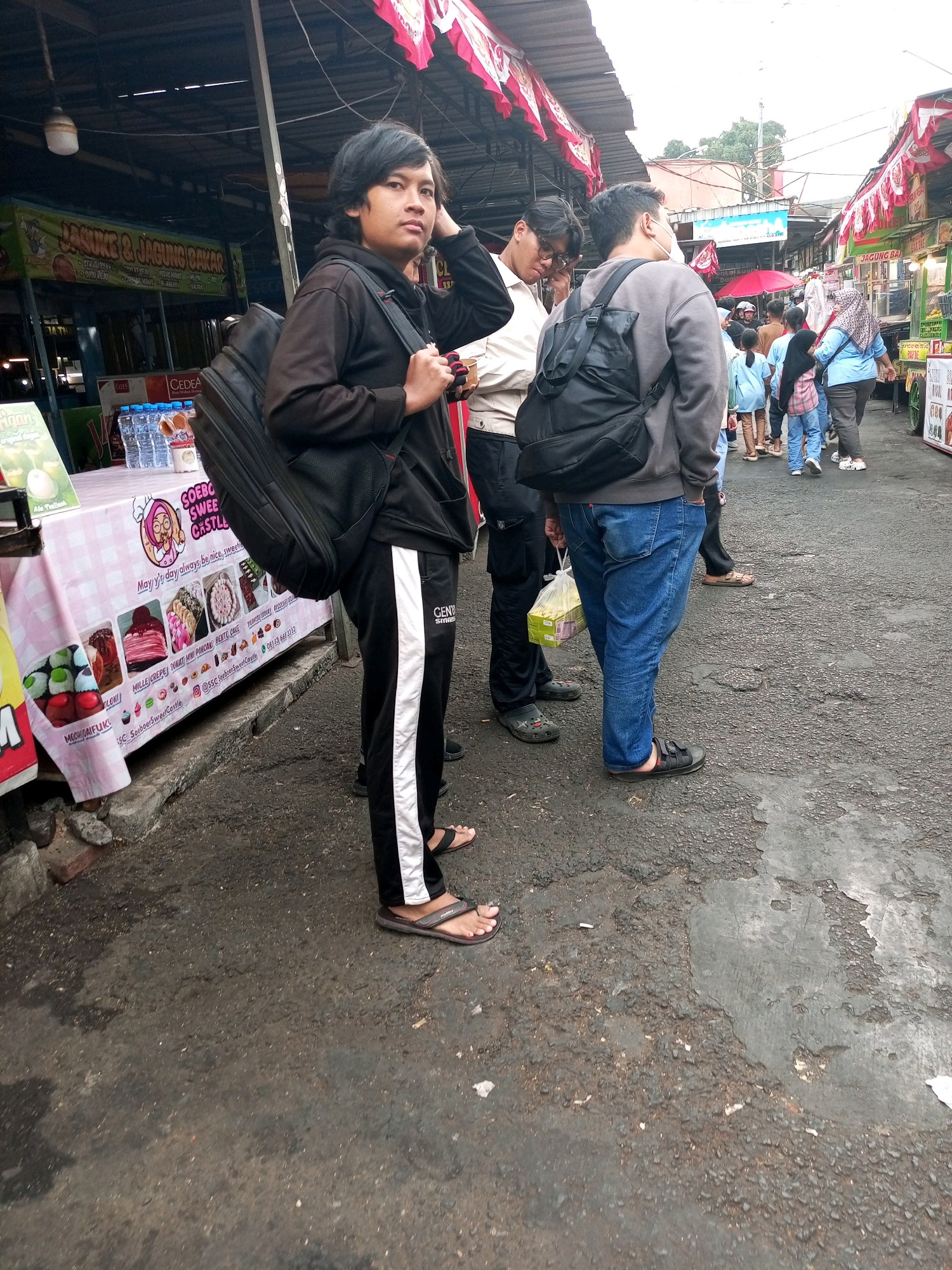 In Batu, Indonesia, a street market scene unfolds. A young man in a black hoodie and track pants, carrying a large black backpack, stands in the foreground, looking towards the camera. To his right, another man in a grey sweatshirt and blue jeans holds a yellow plastic bag. Behind them, other individuals are visible, some wearing masks, as they navigate the market. Stalls lining the street display various goods, including bottles of water and food items like cakes and what appears to be sushi. The ground is paved with asphalt, showing signs of wear and tear. Red and white banners hang overhead, suggesting a festive or celebratory occasion. The overall atmosphere appears to be one of casual commerce and public gathering in an outdoor urban setting.