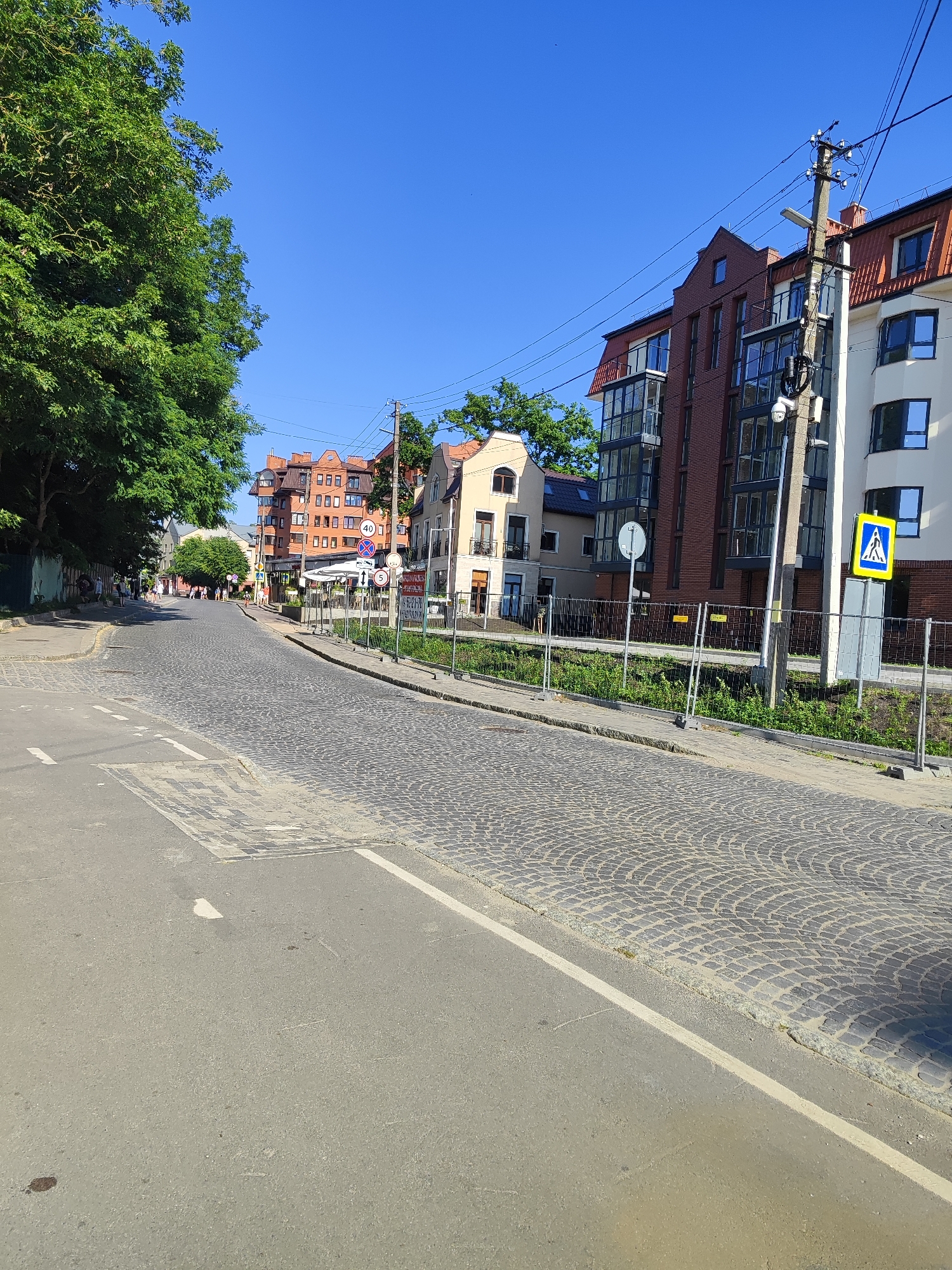 A straight-on, eye-level shot captures a cobblestone street curving to the left in a European-style town. On the left, a large, leafy green tree shades a narrow sidewalk where a few people walk. The right side of the street is lined with multistory buildings, some with red-tiled roofs and brick facades, others painted white. A modern building with large glass windows and balconies is also visible. Temporary fencing runs along the right side of the street, suggesting ongoing construction or maintenance. A traffic light pole with a camera and multiple wires stands prominently near the fenced area. A pedestrian crossing sign is posted on the right, and several other traffic signs, including one indicating a 40 km/h speed limit, are visible. The sky is a clear, vibrant blue, indicating a sunny day. The lighting suggests it is late morning or early afternoon. The scene is tranquil, with no overt activity beyond people walking and the general appearance of an inhabited urban area. The cobblestone pavement, architectural style of the buildings, and the traffic signs hint at a location in Europe, consistent with Pionersky, Russia.