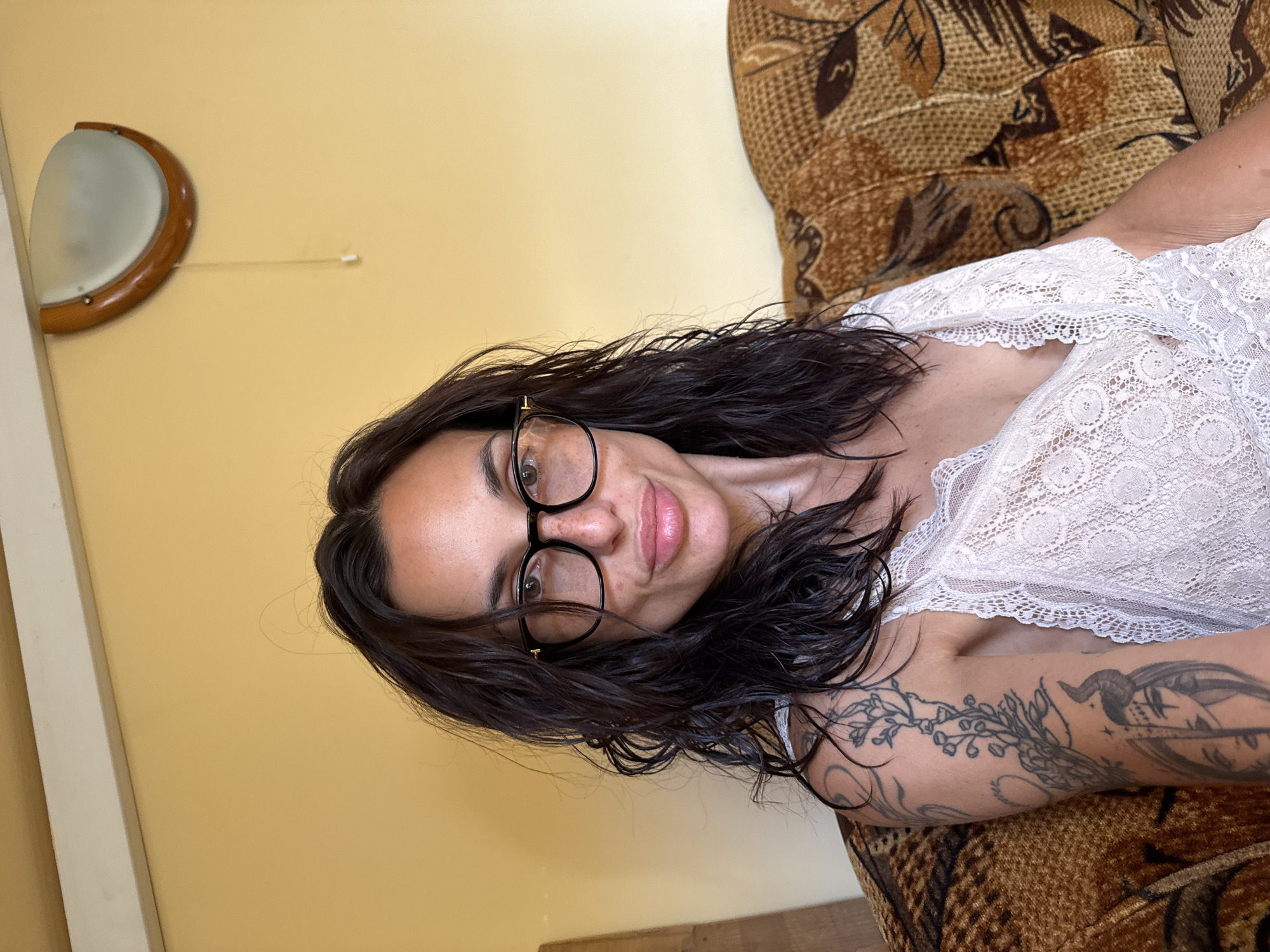 An individual with long, dark, wet hair and glasses is sitting on a couch. They are wearing a white lace top. The individual has tattoos on their arm. The background is a yellow wall with a light fixture.