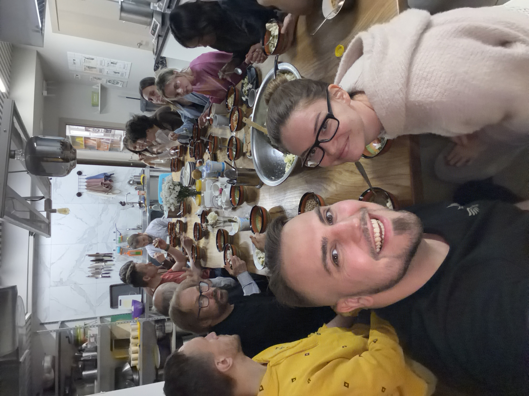 A group of people are gathered around a long wooden table, eating from bowls. The table is set with bowls, plates, utensils, flowers, and water pitchers. The people appear to be engaged in conversation. The location appears to be a commercial kitchen with stainless steel appliances, shelves, and tiled walls. It is not possible to determine if the location is Moscow, Russia, from the image.