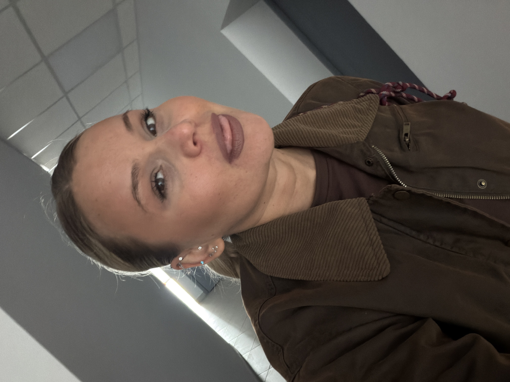 A fair-skinned person is seen from a low angle in a photograph. They are wearing a brown jacket with a corduroy collar and multiple earrings in their left ear. Their hair is pulled back. The person has medium length lashes and their lipstick is a neutral tone. The background suggests a contemporary office or public building with a grid pattern on the ceiling and a neutral-colored wall. There are no indications that this image was taken in Sarapul, Russia.