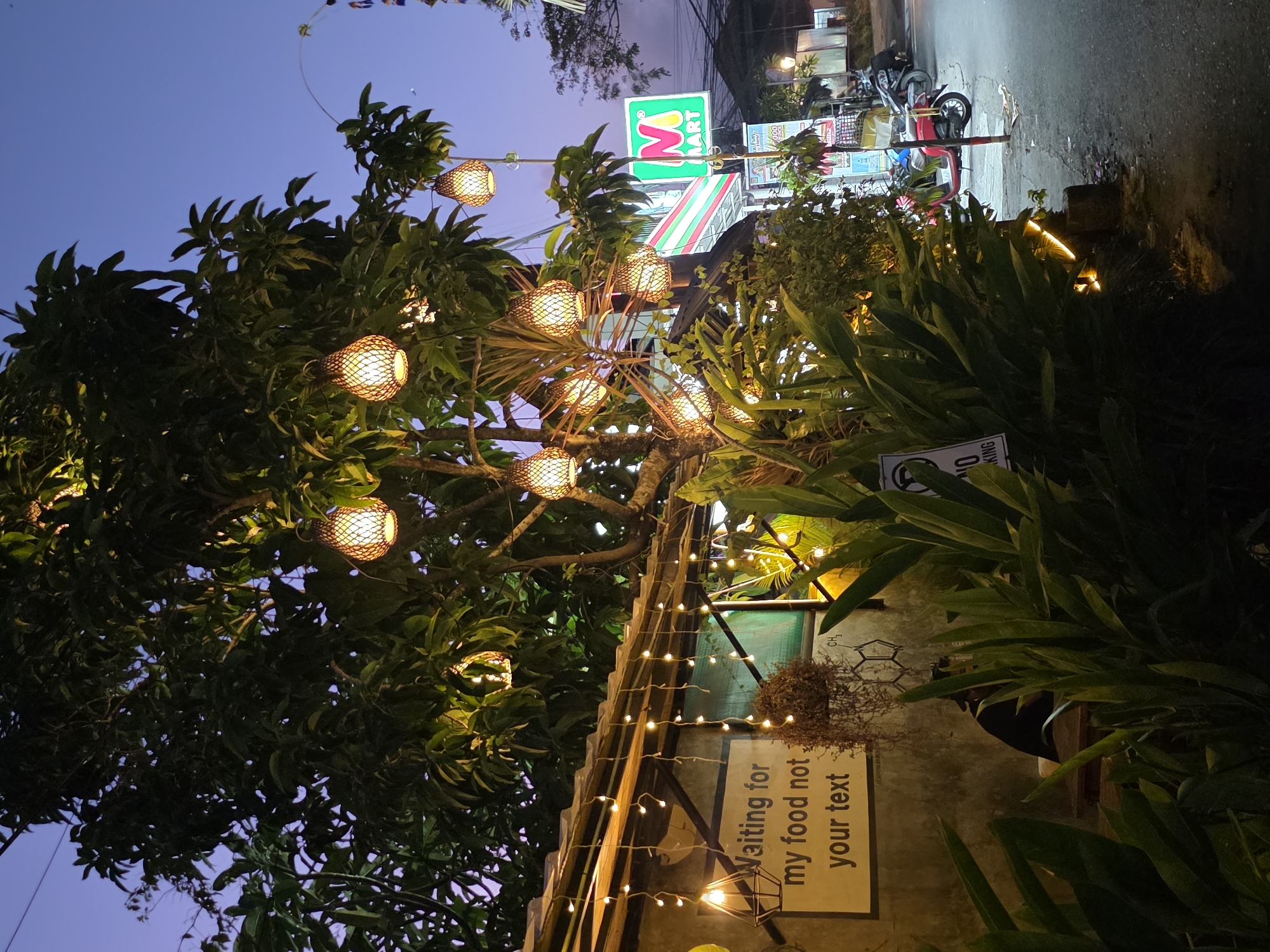 An outdoor scene at dusk in North Kuta, Indonesia. A tree decorated with numerous lantern-like lights is prominent in the foreground, casting a warm glow. Strings of smaller lights also adorn a nearby roofline. A sign on a textured wall reads "Waiting for my food not your text." In the background, a "M MART" convenience store sign is visible, along with parked motorcycles and scooters. The sky is a deep twilight blue.