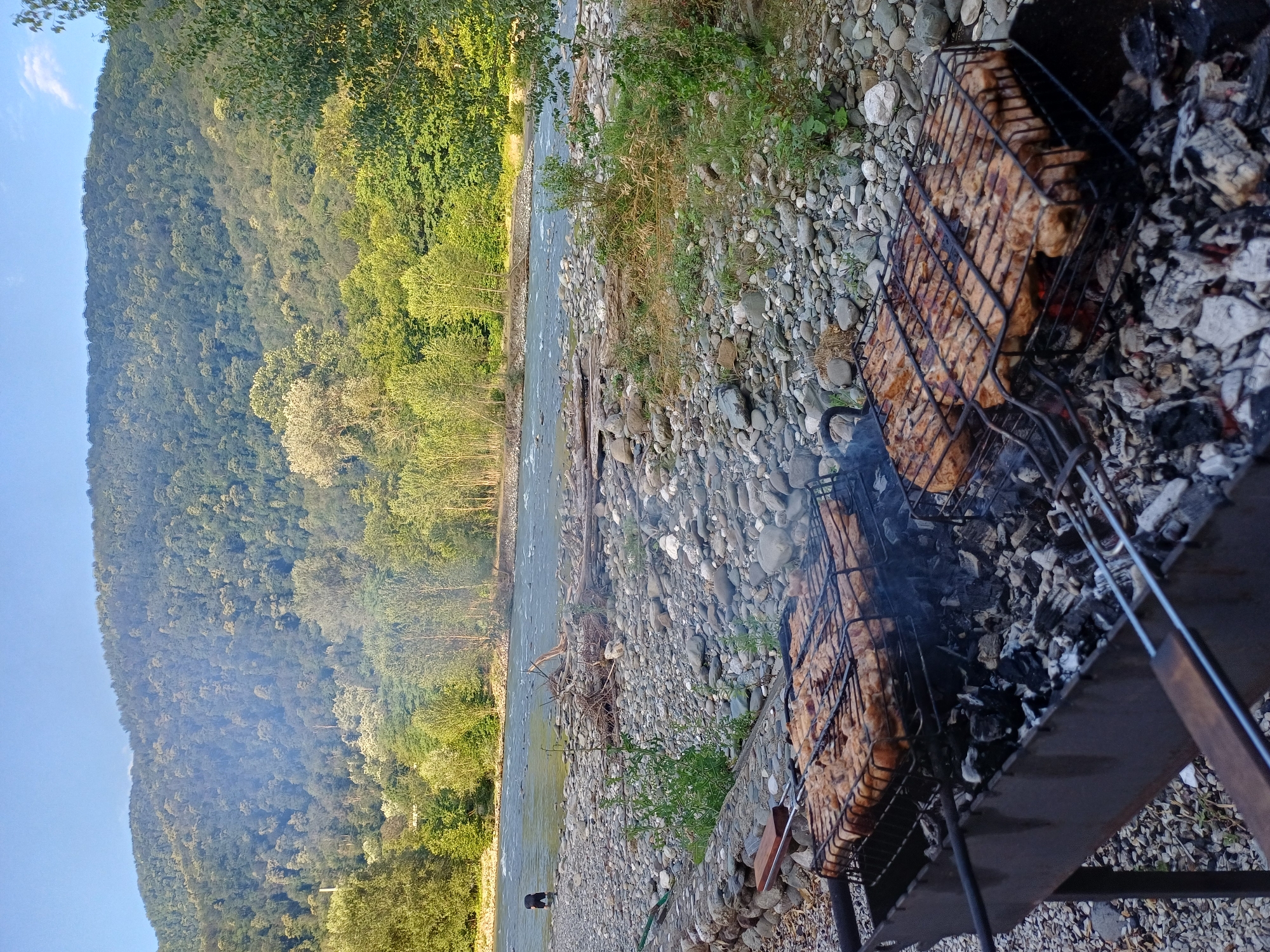 The image depicts an outdoor barbecue scene beside a river, with a dense forest covering a hillside in the background. In the foreground, a metal barbecue grill contains two racks of seasoned meat being cooked over glowing coals. Smoke rises from the grill, indicating the cooking process is underway. The area surrounding the grill is covered in pebbles and rocks. To the left, a small section of a stone wall is visible. Further back, a wide river flows, with a person standing in the water in the middle ground on the left side of the frame. The riverbank is also lined with pebbles and scattered driftwood. The background features a lush green forest that extends up the slope of a mountain, under a clear blue sky. The overall scene suggests a recreational activity in a natural, mountainous environment, likely near the city of Monastyr, Russia, given the context.
