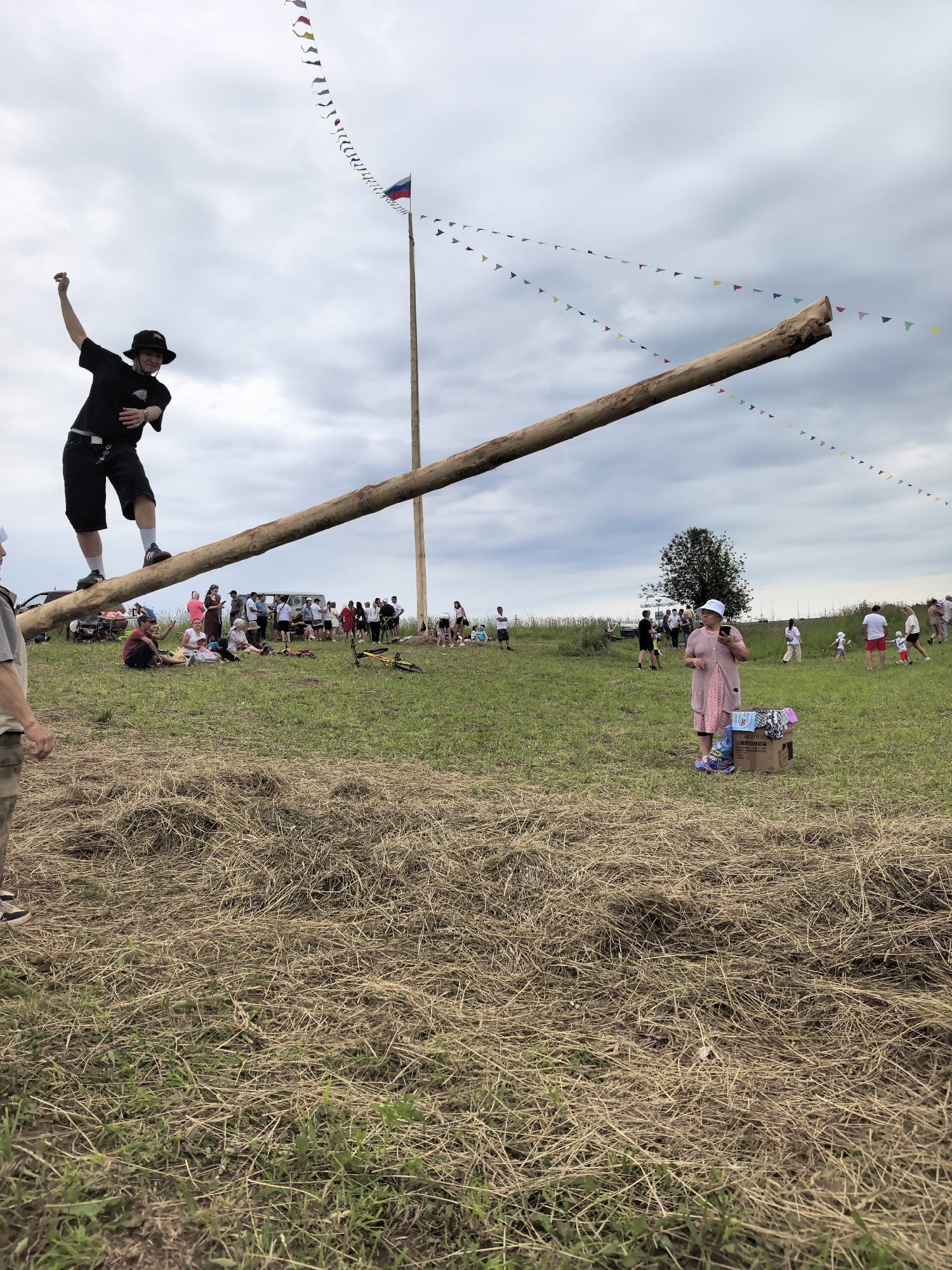 The image depicts an outdoor event, likely a festival or competition, taking place in a grassy field in Koyanovo, Russia, under a cloudy sky. A long, thick wooden log is positioned diagonally across the frame, with one end resting on the ground and the other elevated, supported by a tall wooden pole. A young man wearing a black t-shirt, black shorts, white socks, and a black hat is balancing precariously on the log, with his left arm raised. He is facing towards the left of the frame.
In the background, a crowd of people is gathered, some sitting on the grass and others standing. A tall pole with a Russian flag is visible in the distance. Colorful triangular flags are strung between poles and appear to be decorations for the event. Several vehicles, including a white van and a red car, are parked in the background.
To the right of the frame, an older woman wearing a pink dress and a white hat stands near a cardboard box filled with items, possibly prizes or supplies. She is holding a smartphone in her hand. Further in the background on the right, other people, including children, are visible mingling. The foreground of the image is covered with dry hay and green grass, suggesting a rural or semi-rural setting. A bicycle lies on its side in the middle ground.