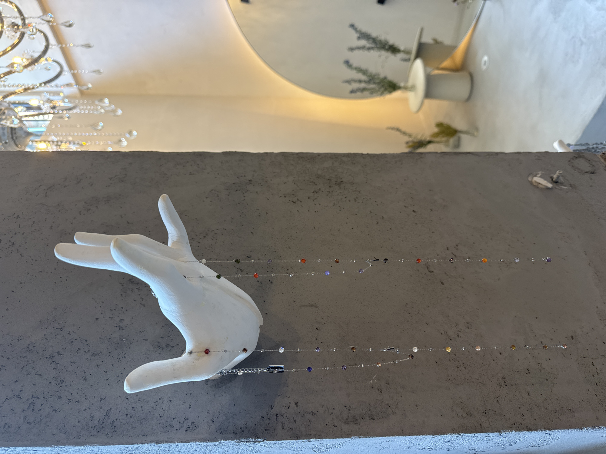 The image shows a white sculpture of a hand mounted on a grey concrete wall. Two delicate necklaces with colorful beads hang from the fingers of the sculpture. In the background, a blurred scene reveals a chandelier with crystal drops, a circular mirror with illuminated edges, and a small table with vases of greenery. The overall aesthetic suggests an interior space, possibly a boutique or a gallery, with a modern and elegant design. The location is identified as Sofia, Bulgaria.