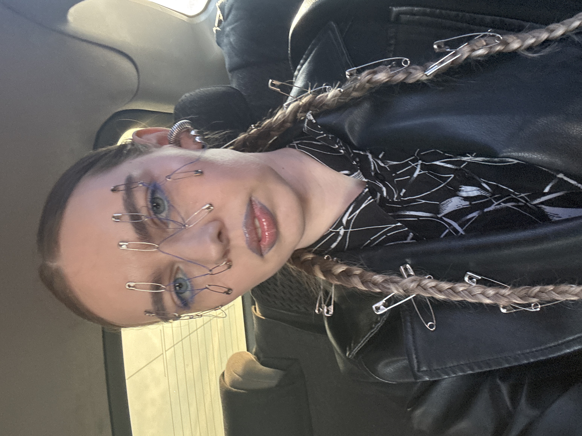 A person is shown in a close-up shot. The person's hair is styled into braids adorned with safety pins. More safety pins are attached to the person's face with blue string or thread. The person is wearing a black leather jacket. The background shows a vehicle interior and, indistinctly, a building through the car window. This image was taken in the city of Krasnoyarsk, Russia.