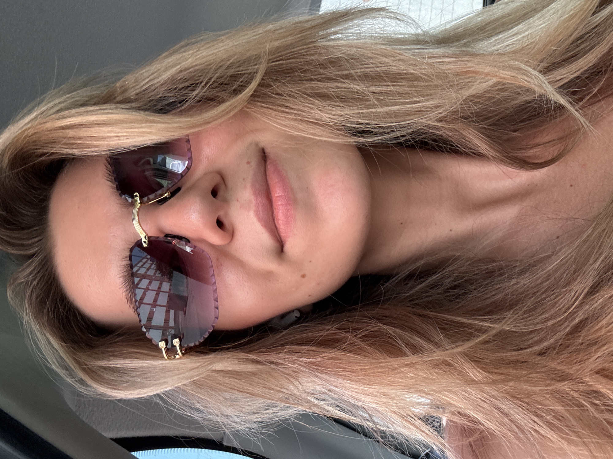 A close-up, medium selfie of a young Caucasian woman with blonde hair and fair skin is captured from a slightly upward angle. She is wearing sunglasses with a faceted, heart-shaped lens and gold frames, and appears to be in a vehicle, possibly a car, indicated by the background which shows interior elements and a window reflecting a building with square windows. The woman is looking directly at the camera with a slight smile. She has a dimple on her right cheek and a small mole below her left eye and on her neck. She is also wearing a small white earbud in her right ear. The overall lighting suggests it is daytime.