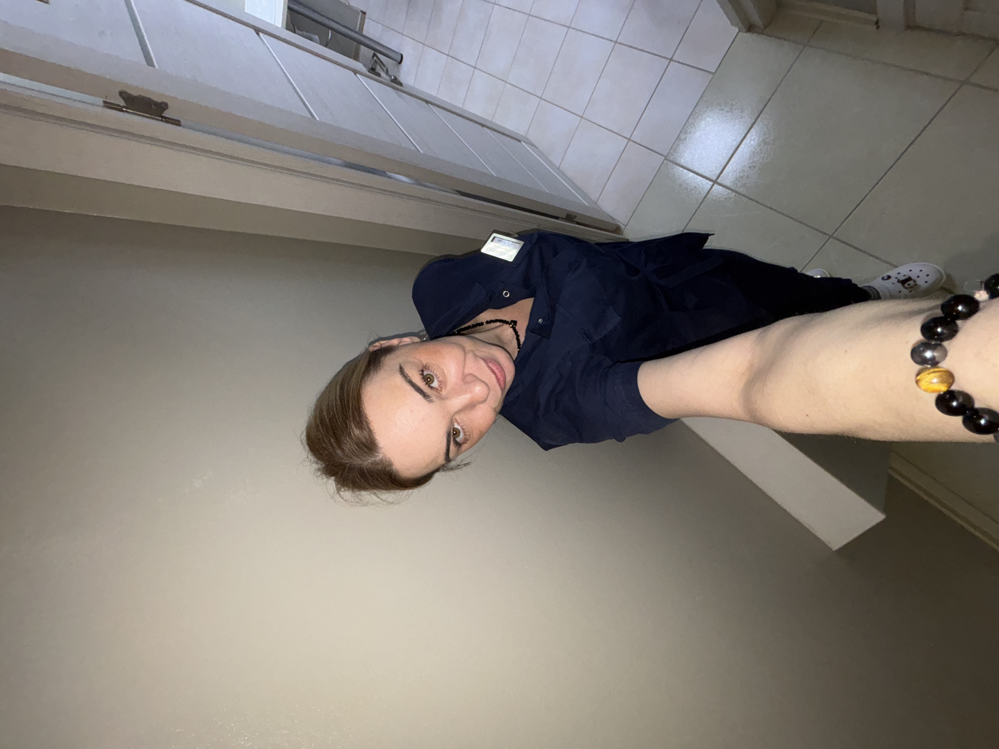 A selfie taken in what appears to be a hallway. The person is wearing a dark blue shirt, a beaded necklace, and a bracelet with dark and amber beads. They are wearing socks with designs and white shoes. The background includes a light-colored wall, a door, and tiled flooring. The exact location in Unknown, Russia, cannot be determined.