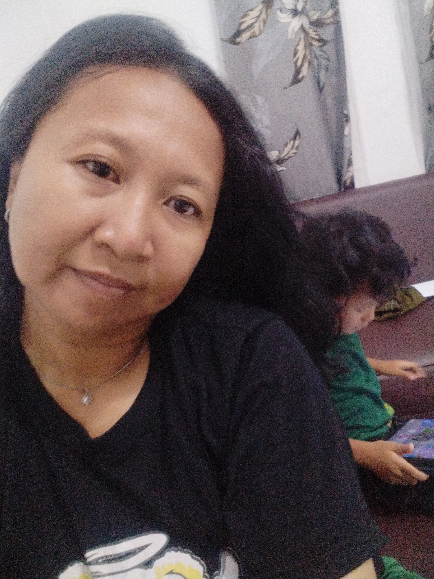 In an indoor setting, likely a living room in Karangsentosa, Indonesia, a woman is captured in a selfie. She has long, dark hair and is wearing a black t-shirt with a delicate necklace. Her expression is relaxed, and she appears to be looking slightly off-camera. Behind her, a child with dark, curly hair is seated on a brown couch and is engaged with a tablet, holding it with their hands. The background features a curtain with a floral pattern. The lighting suggests a moderately lit room, possibly during the daytime.