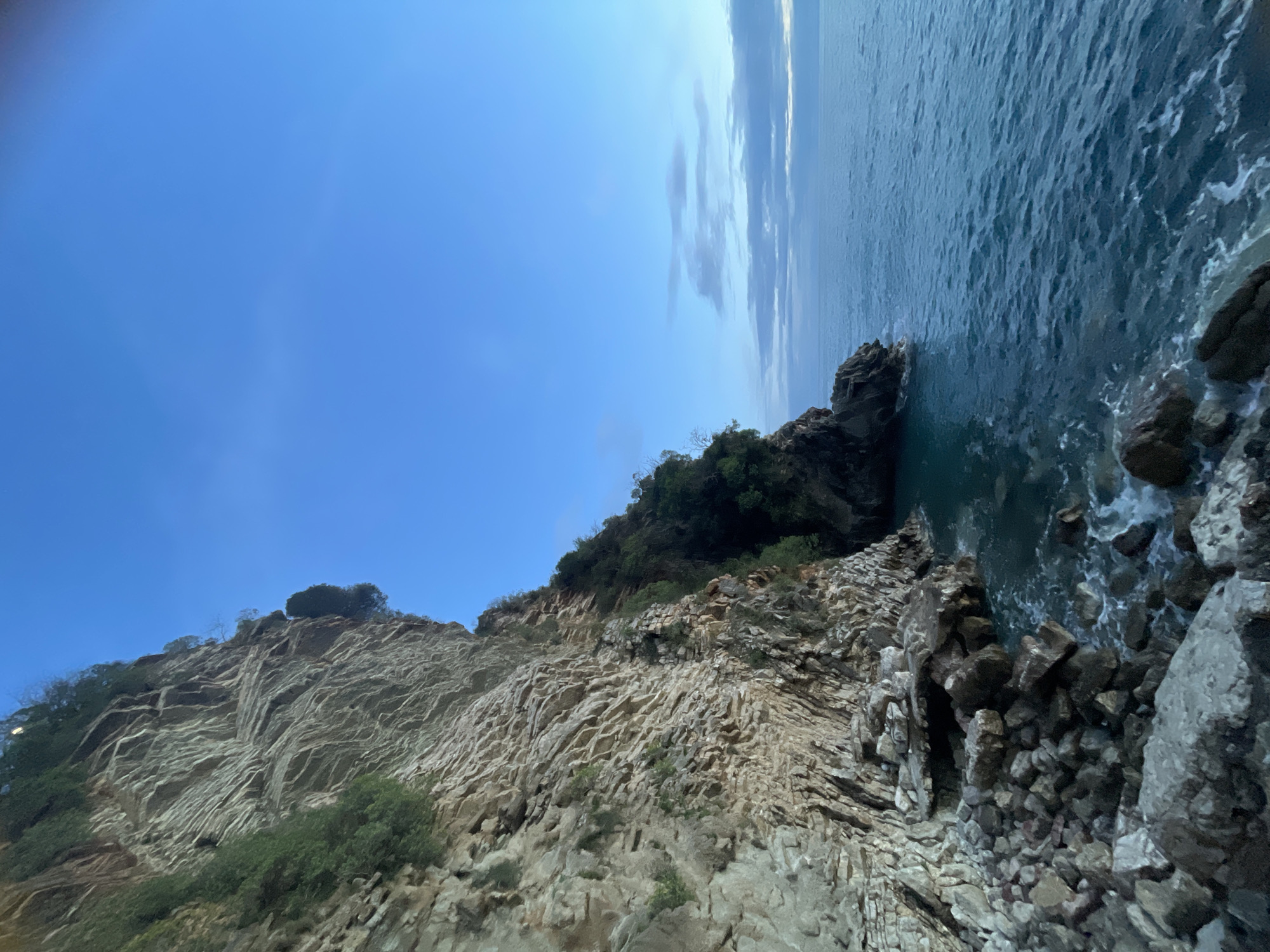 The image displays a coastal landscape in Rafailovići, Montenegro, presenting a large rock cliff meeting the sea. The left two-thirds of the frame are dominated by a towering, stratified cliff face, composed of light grey, beige, and brown rock with visible vertical fractures and horizontal layering. Green vegetation, consisting of shrubs and small trees, clings to crevices and along the upper portions of the cliff. At the base of the cliff, a rugged shore comprises numerous dark grey and brown boulders and rocks, some partially submerged in the water where small waves break, generating white foam. The right side of the image features the sea, exhibiting deep blue-green water with a textured surface indicating gentle wave activity. The sea extends to a visible horizon line. Above the horizon, the sky is clear and light blue, transitioning to a paler hue with scattered, wispy grey clouds. The overall lighting suggests either dawn or dusk. No human figures or artificial structures are present in the scene.