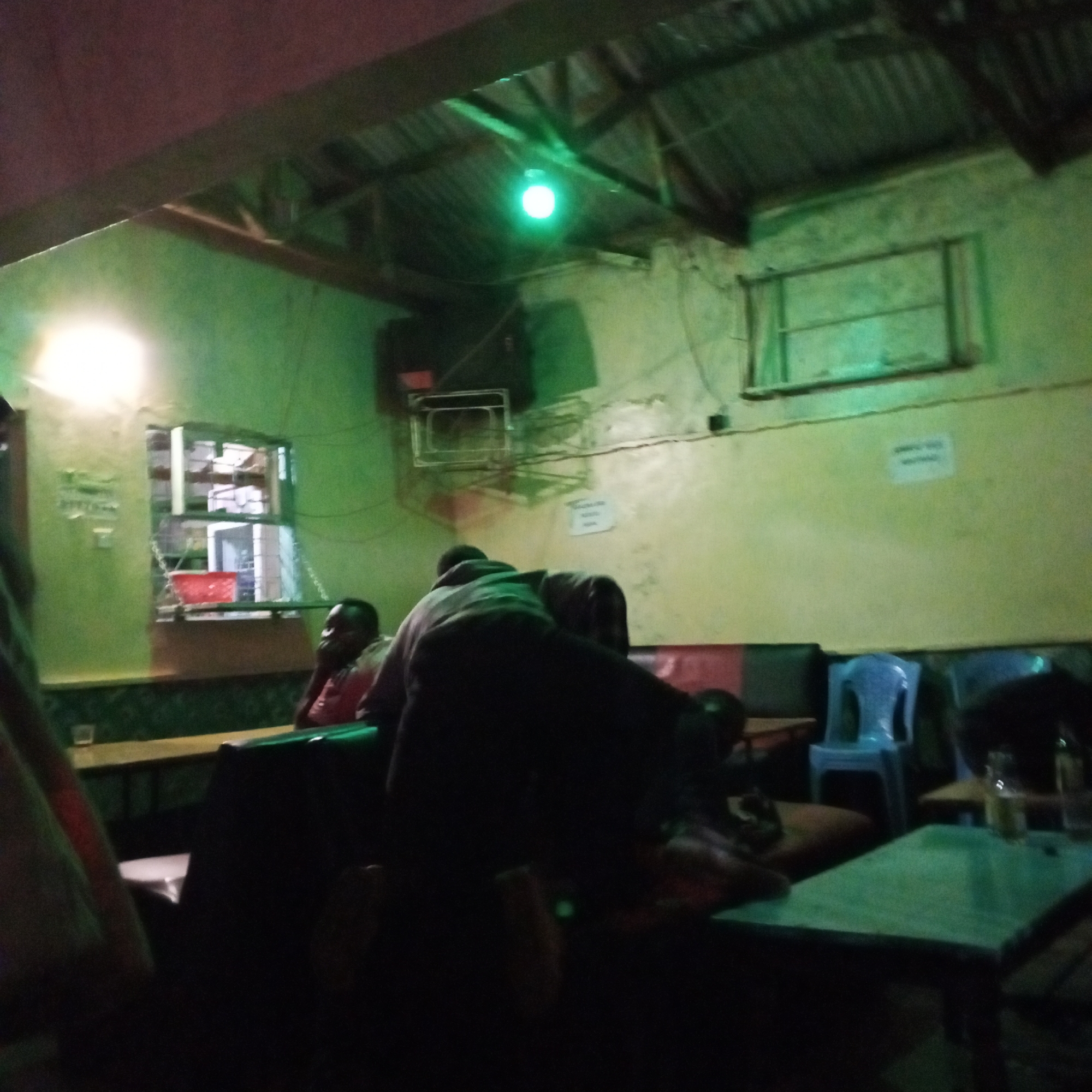 The image depicts the interior of a dimly lit establishment, possibly a bar or restaurant, in Borabu Chitago ward, Kenya. The walls are a faded greenish-yellow color, and the ceiling appears to be corrugated metal. The lighting is provided by a bright white light and a green light fixture in the ceiling. Several people are present, some seated at tables and others standing. Furniture includes wooden tables, a padded booth-style seat against the wall, and a blue plastic chair. There are windows on the left side of the image. An electronic device is attached to the wall. A sign is on the wall next to the electronic device. Some drinking glasses and bottles are on the tables. The overall environment is dark and possibly smoky.